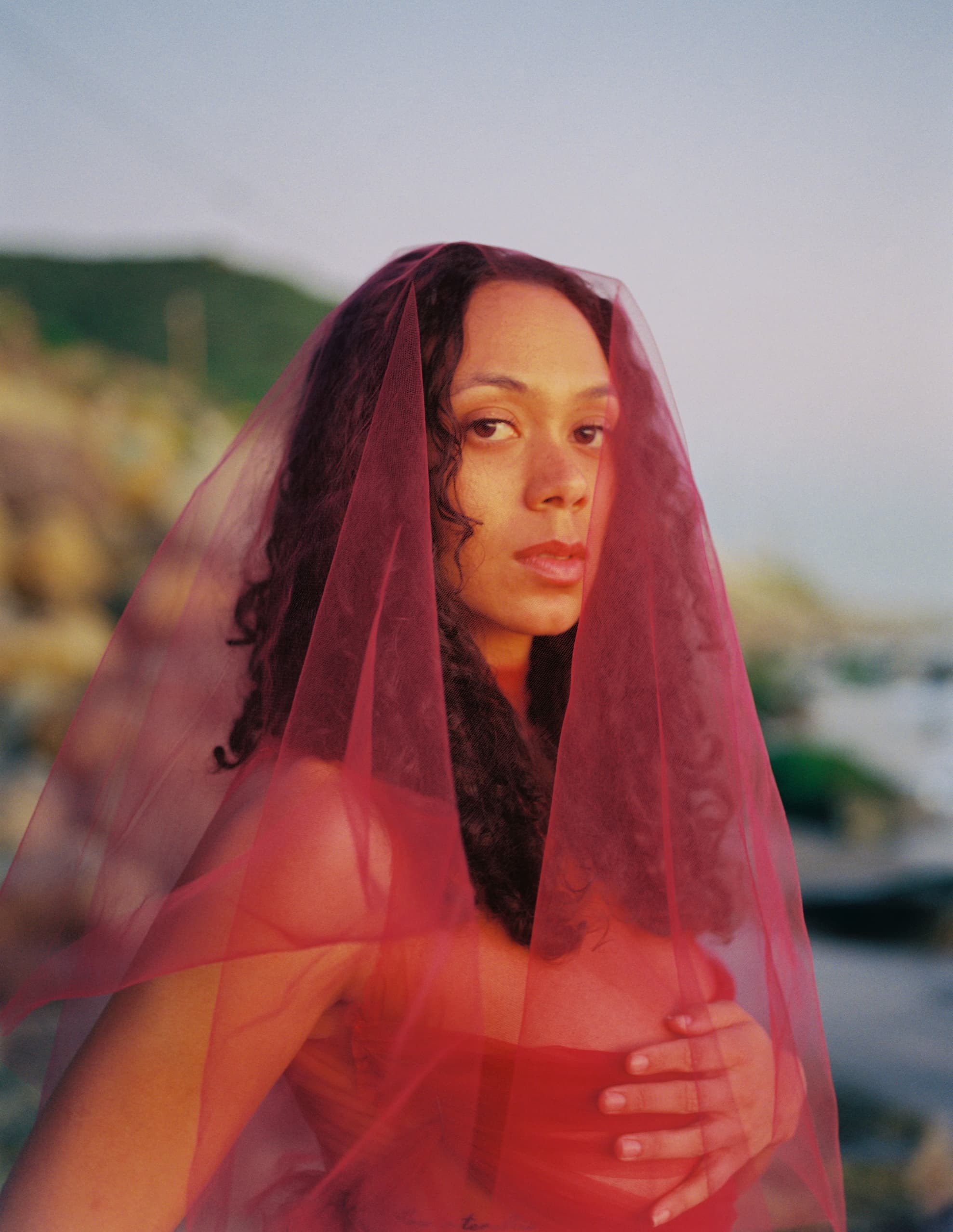 Woman in red veil at sunset, standing outdoors with serene expression.