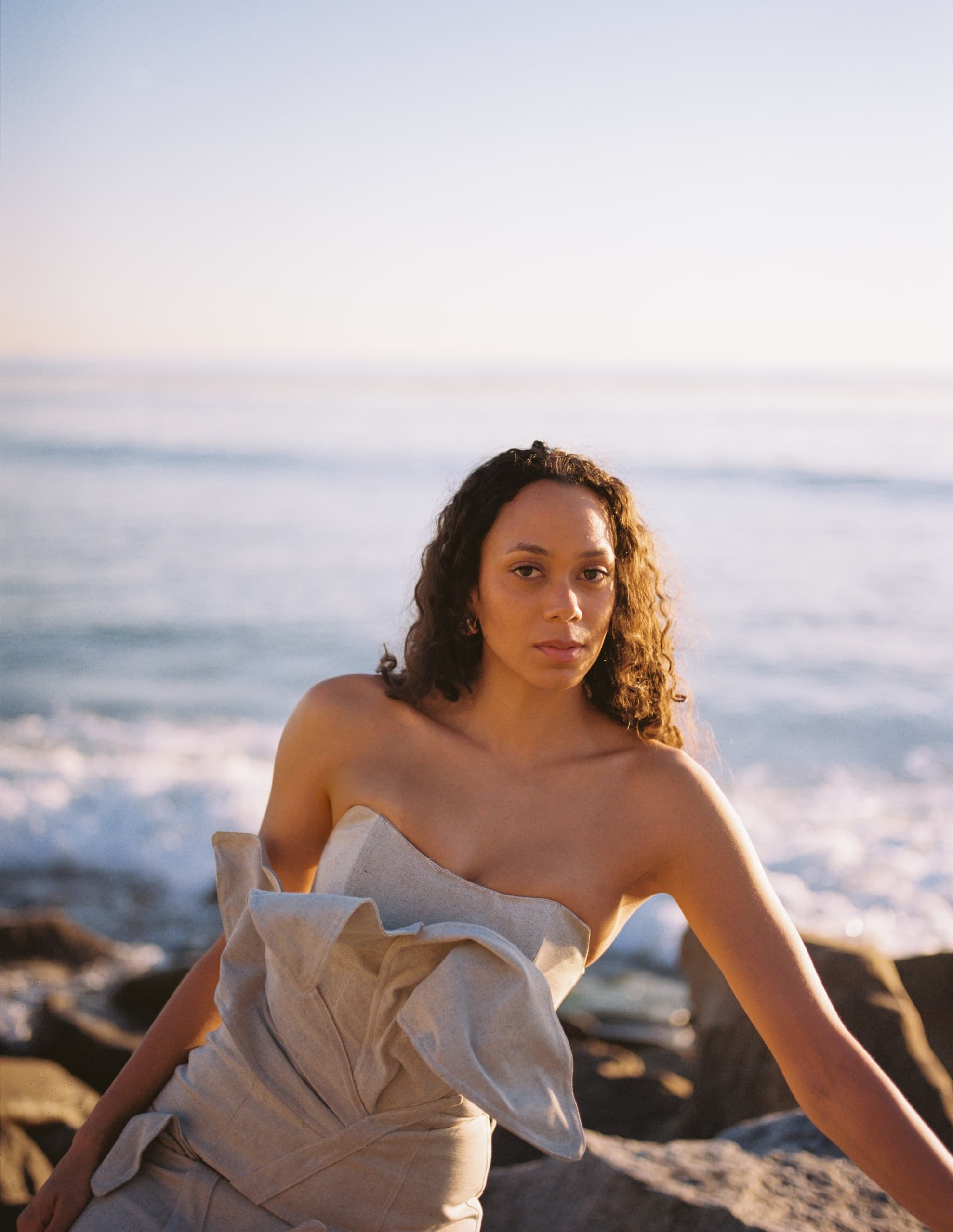 Woman in elegant dress poses by the ocean at sunset.