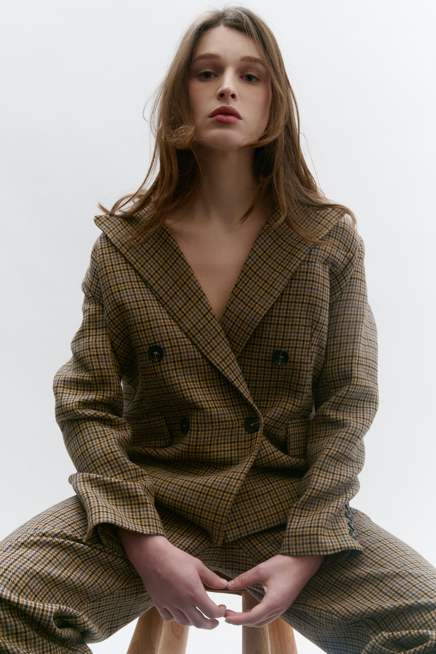 Woman in plaid suit sitting on a stool, posing confidently against a neutral background.
