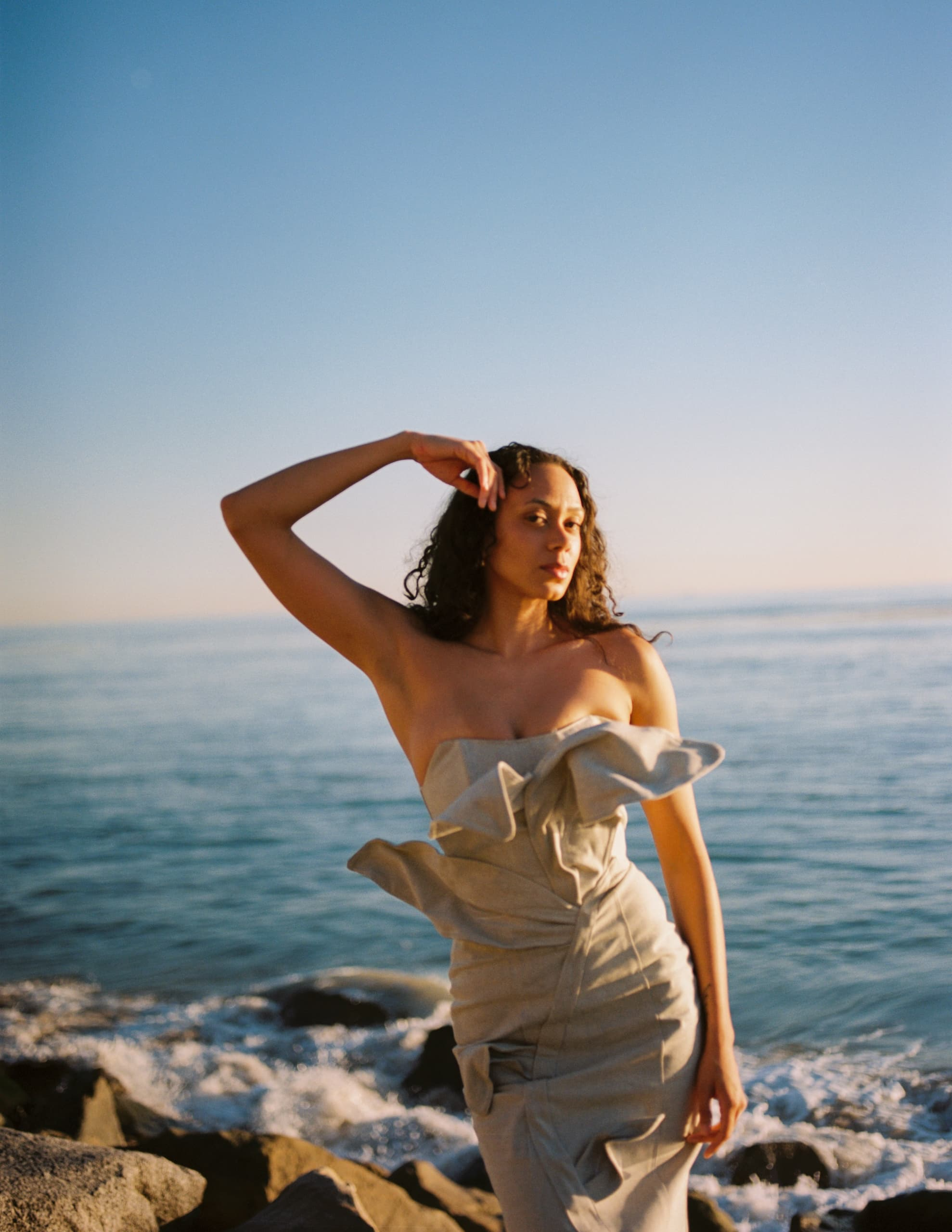 Woman in artistic dress poses by the ocean at sunset.