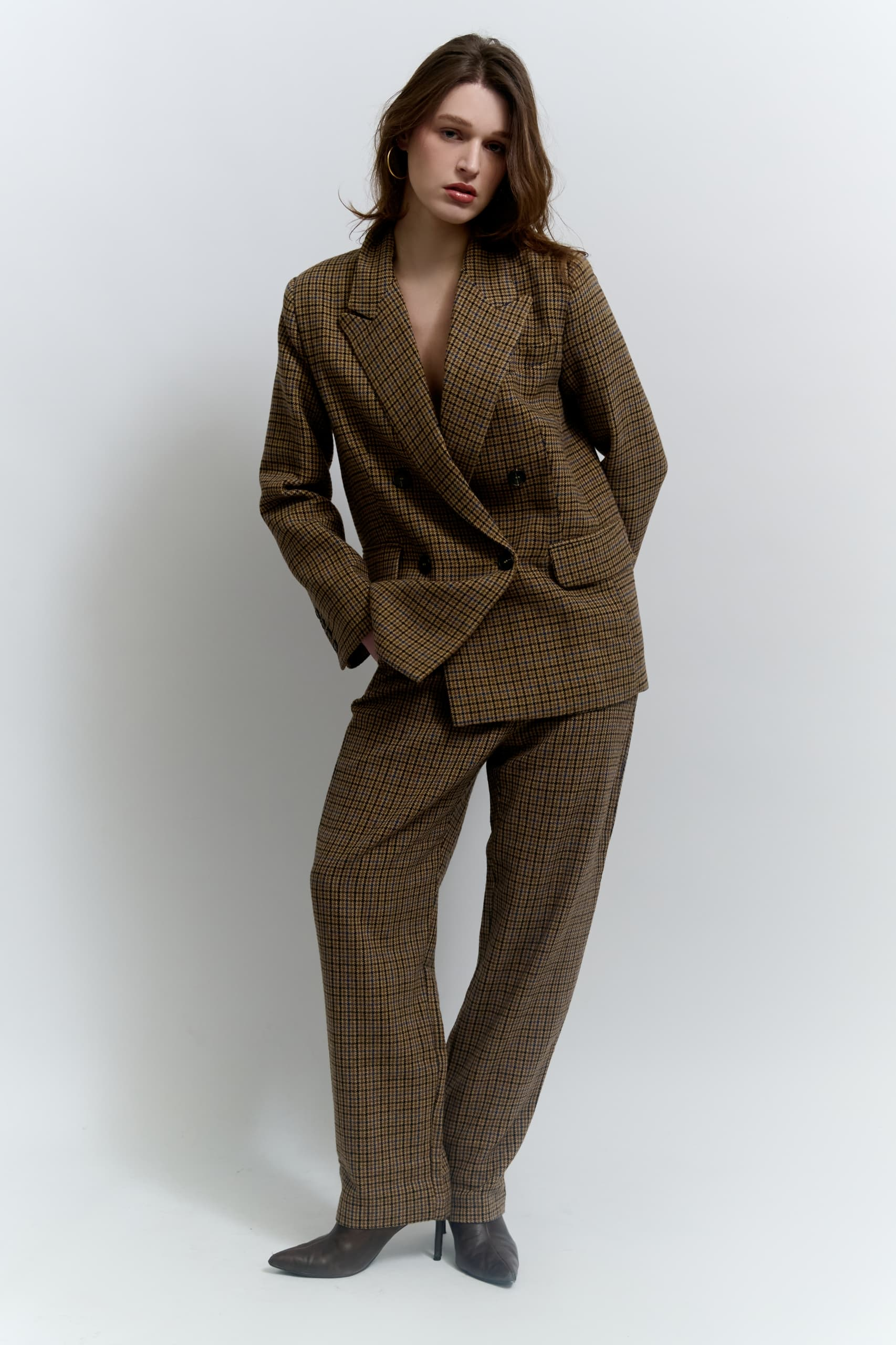 Woman in stylish plaid suit with high heels, posing confidently against neutral background.