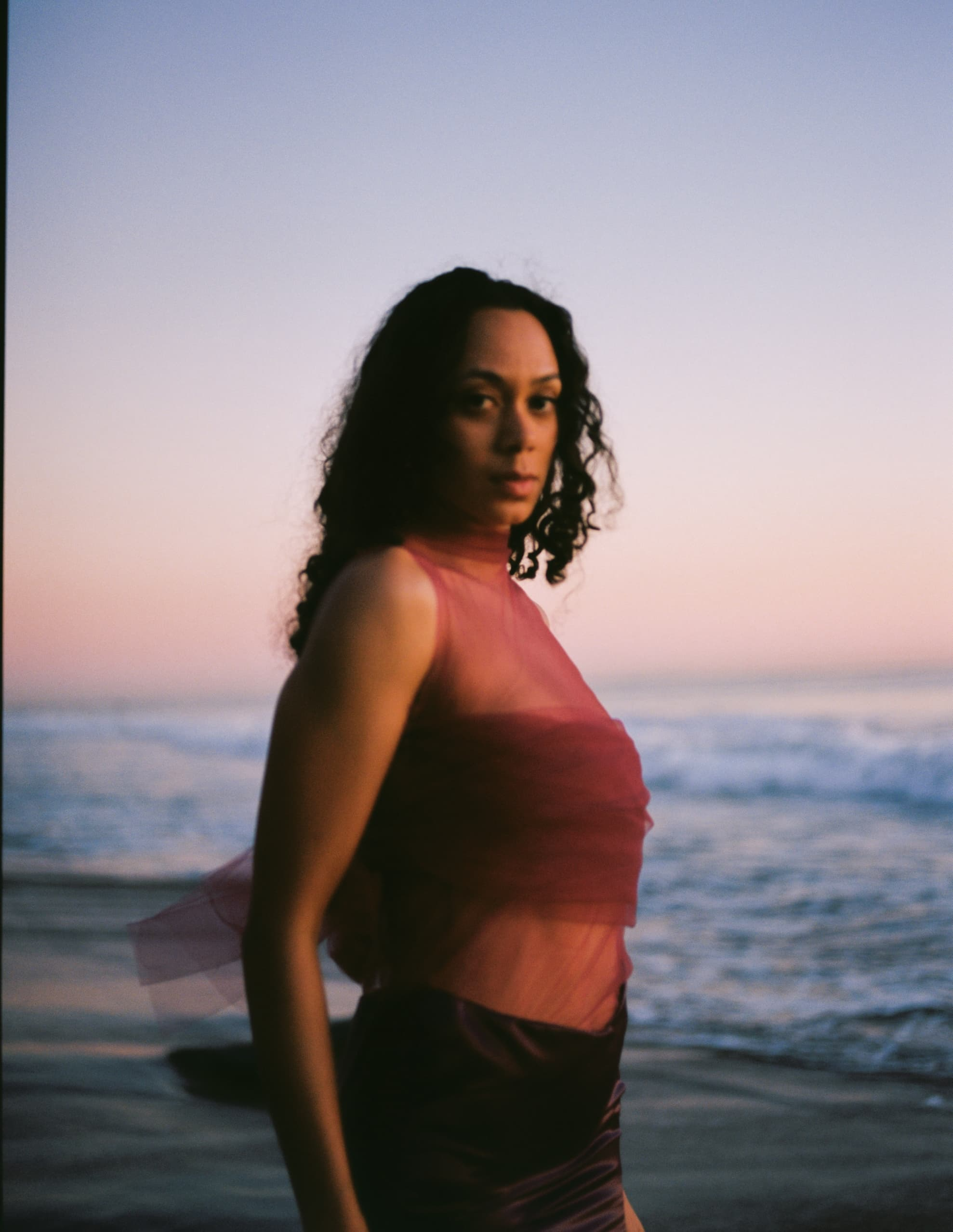 Woman in red dress standing on beach at sunset with ocean backdrop.
