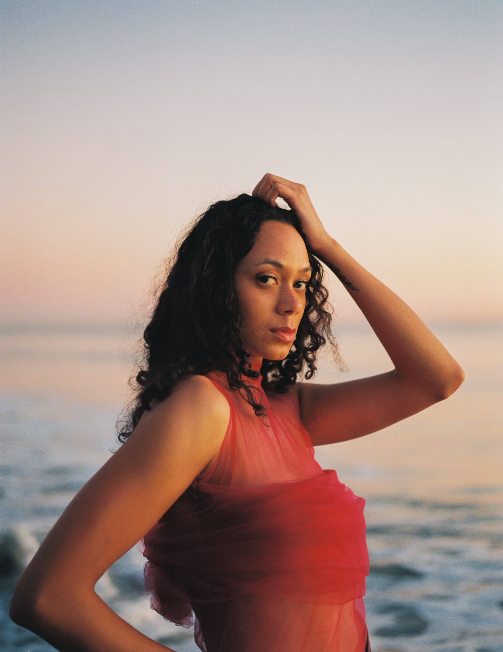 Woman in red dress by the ocean at sunset, with contemplative expression.