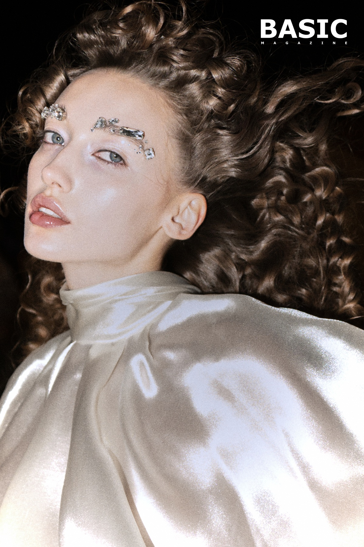 Fashion model with curly hair and jeweled eyebrows in satiny attire.