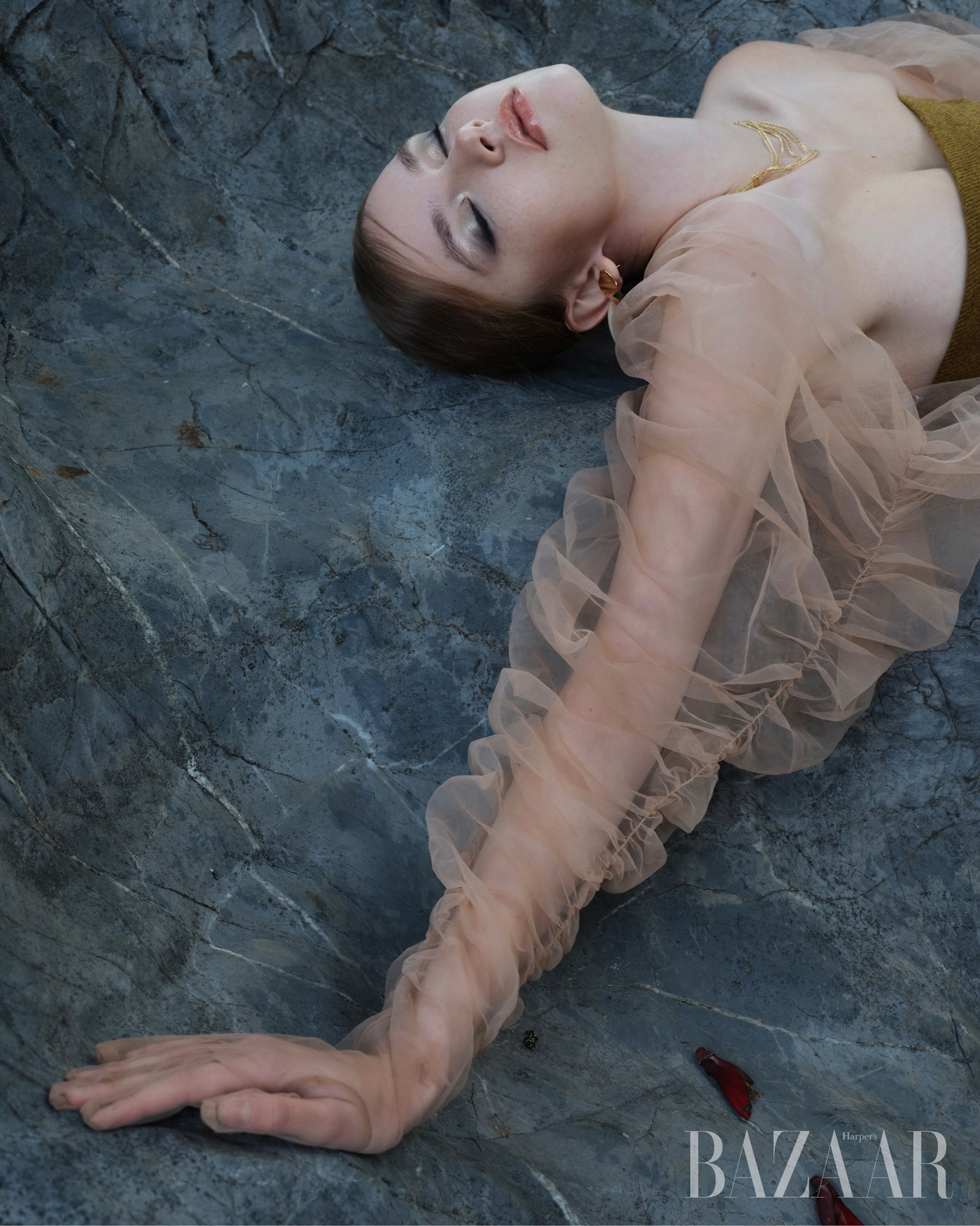 Elegant woman on stone, wearing sheer dress, serene pose, fashion photography.