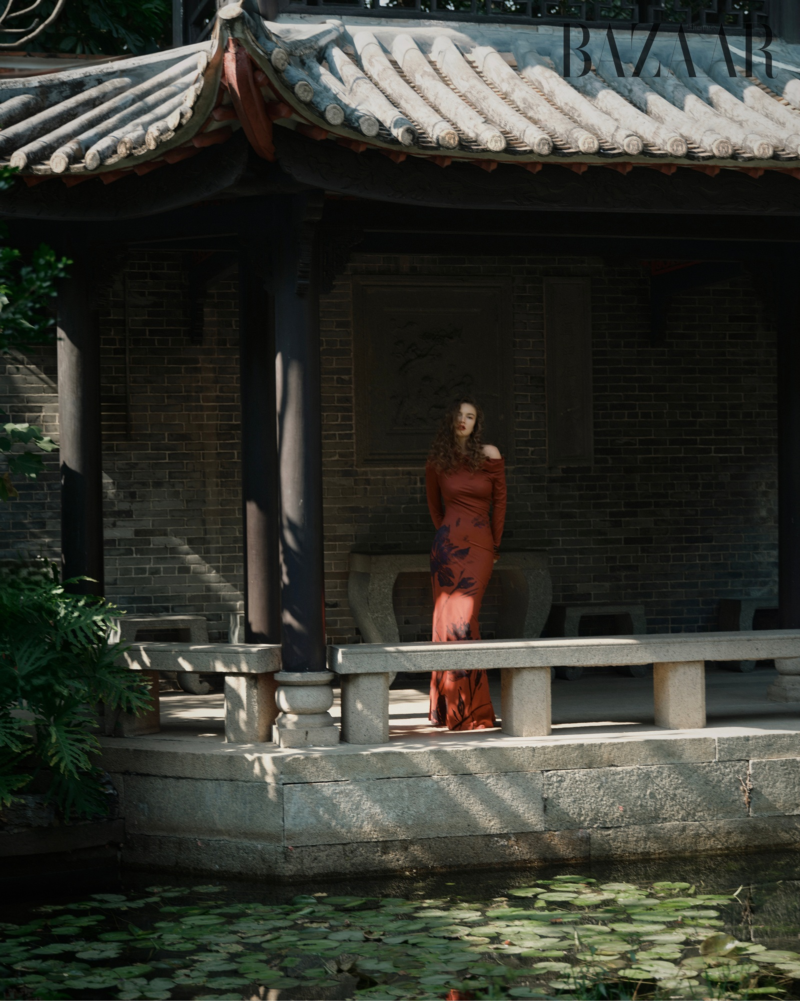A woman in a red dress stands in a serene Asian garden pavilion by a pond.