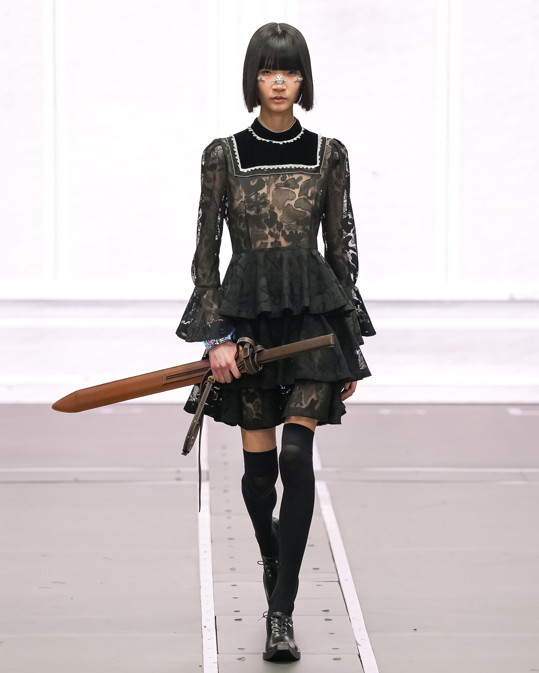 Model on runway in black lace dress holding a sword accessory.