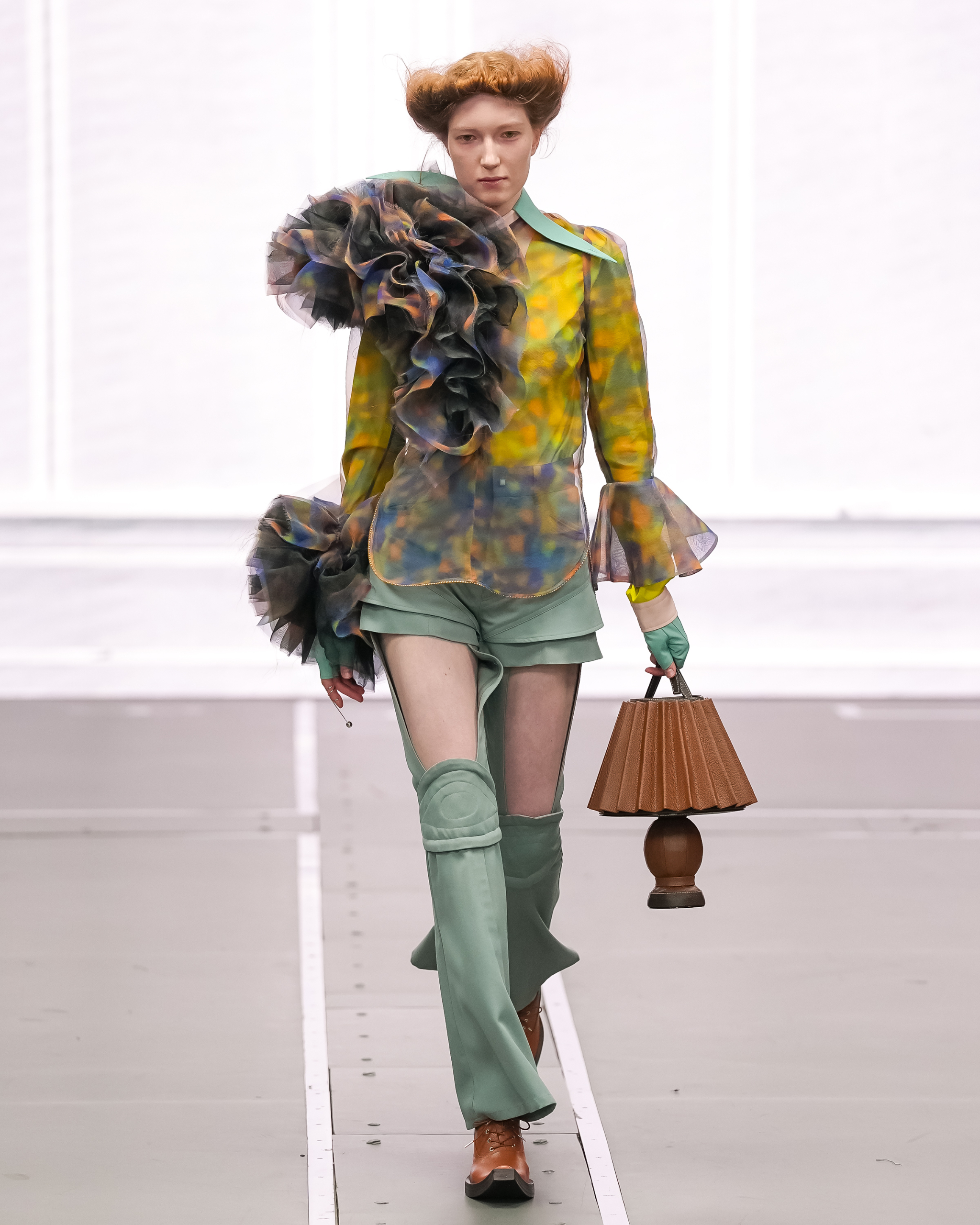 Model in colorful avant-garde outfit on runway, holding a unique lamp-shaped handbag.