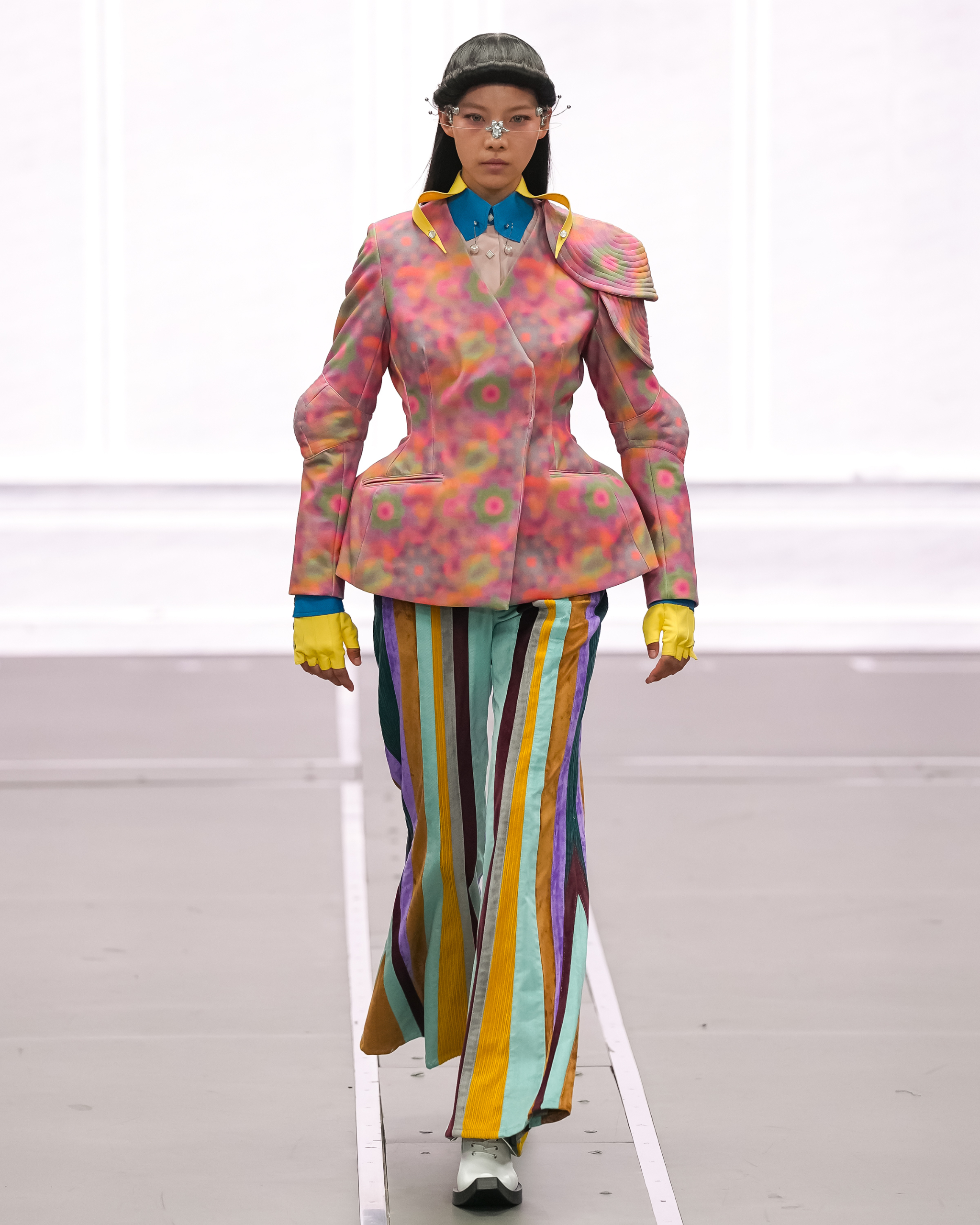 Model in vibrant outfit with striped pants and floral jacket on runway.