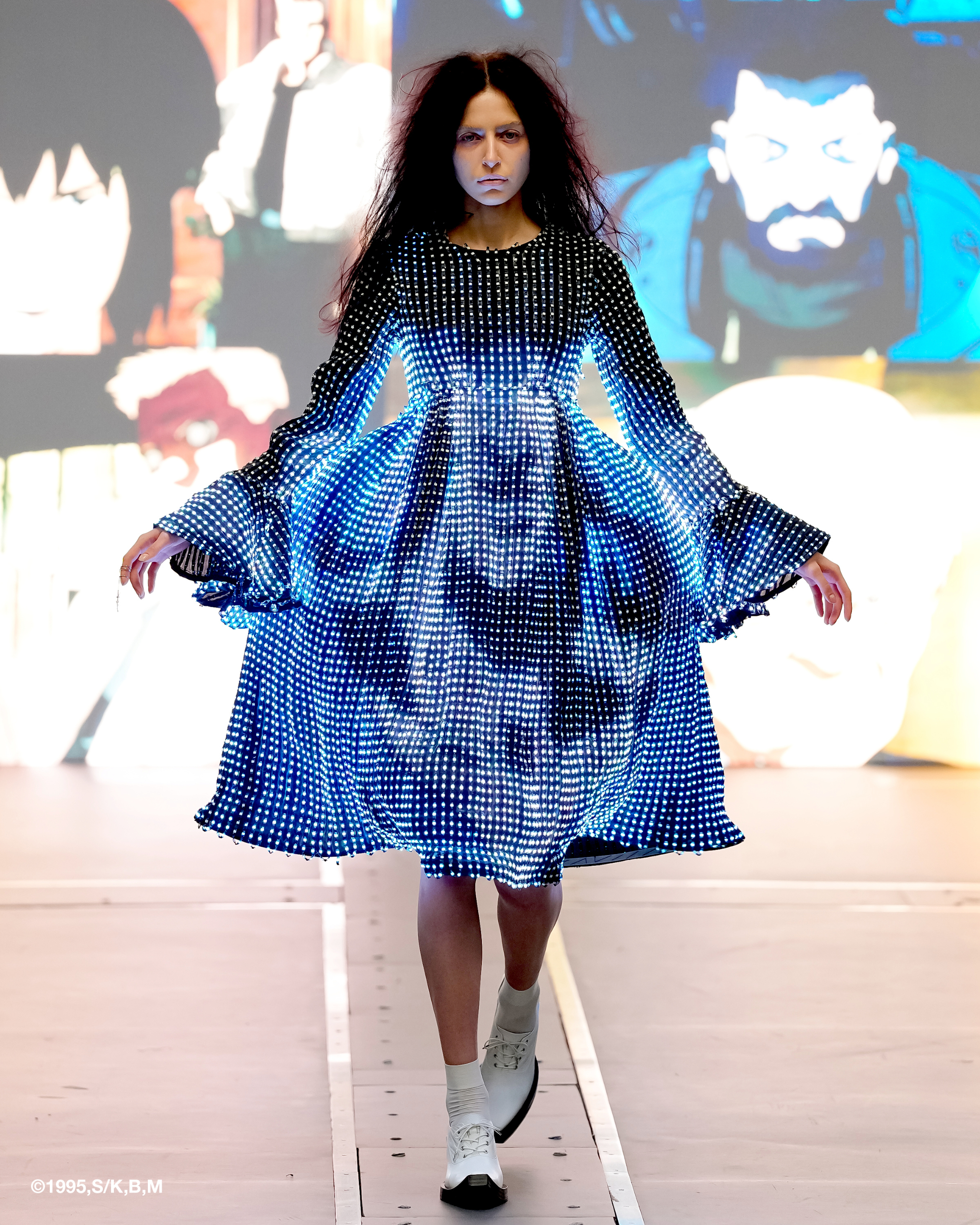 Model in LED dress walks runway with animated backdrop.
