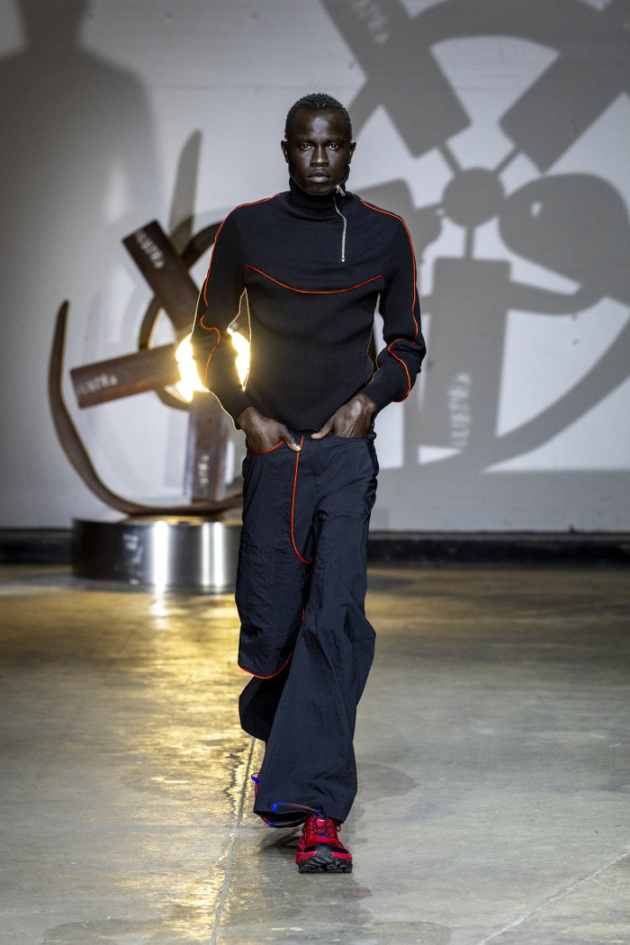 Model in black and red outfit walking runway with artistic backdrop.