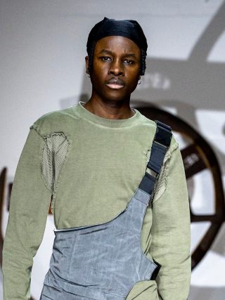 Model in green top and gray outfit on a fashion runway stage.