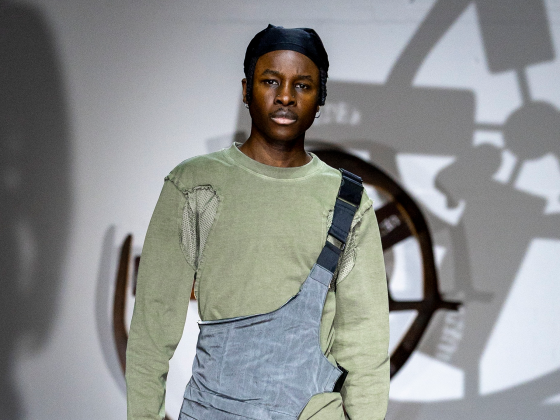 Model in green top and gray outfit on a fashion runway stage.