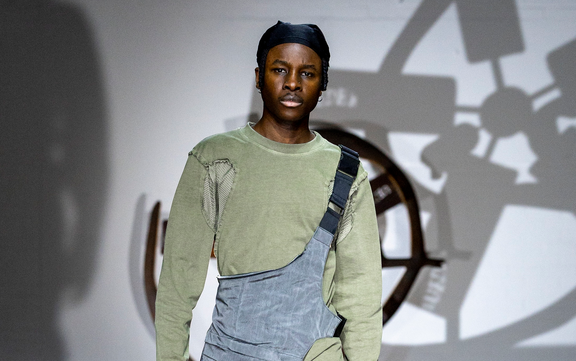 Model in green top and gray outfit on a fashion runway stage.