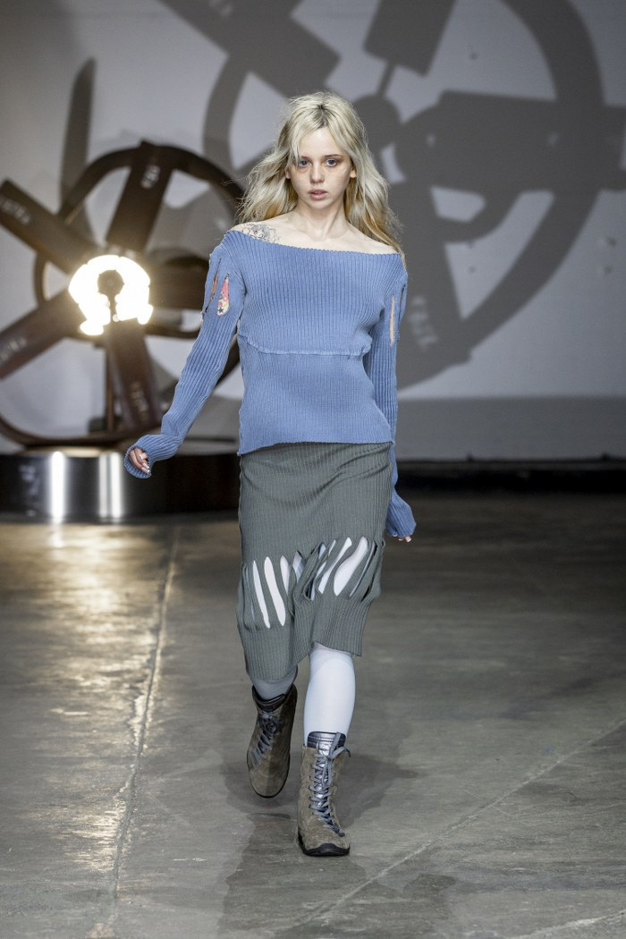 Model in chic fashion on runway, edgy outfit with ripped sweater and skirt.