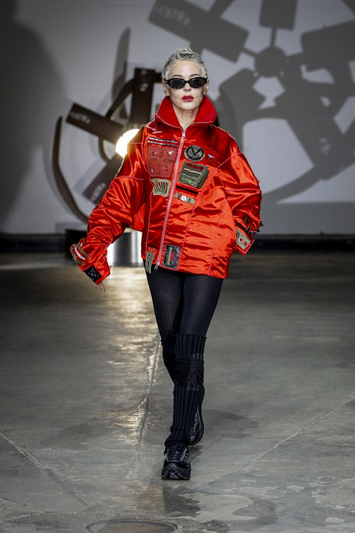 Model in vibrant red jacket and sunglasses on runway.