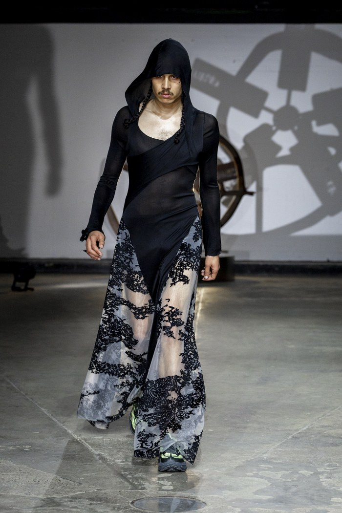 Model in gothic black hooded dress on runway with intricate lace detailing.