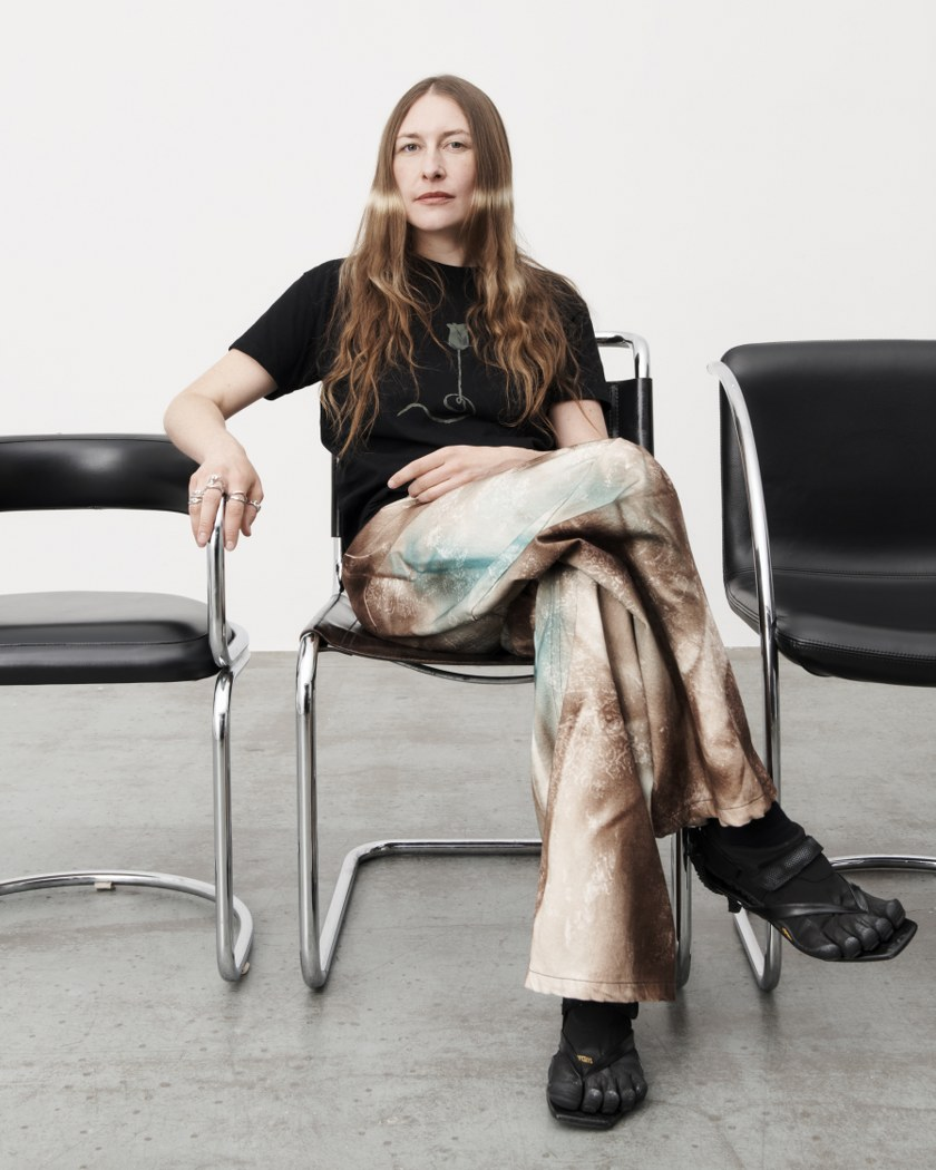 Person sitting in a chair wearing artistic pants and a black shirt with long hair.
