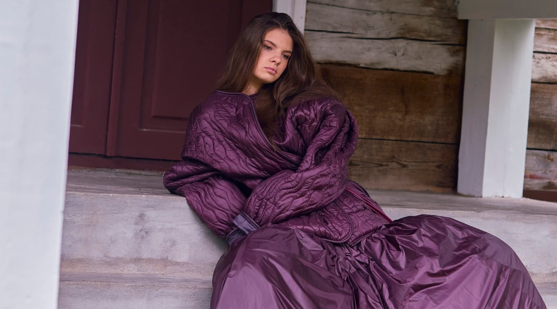 Woman in a purple quilted outfit sitting on steps by a rustic wooden door.