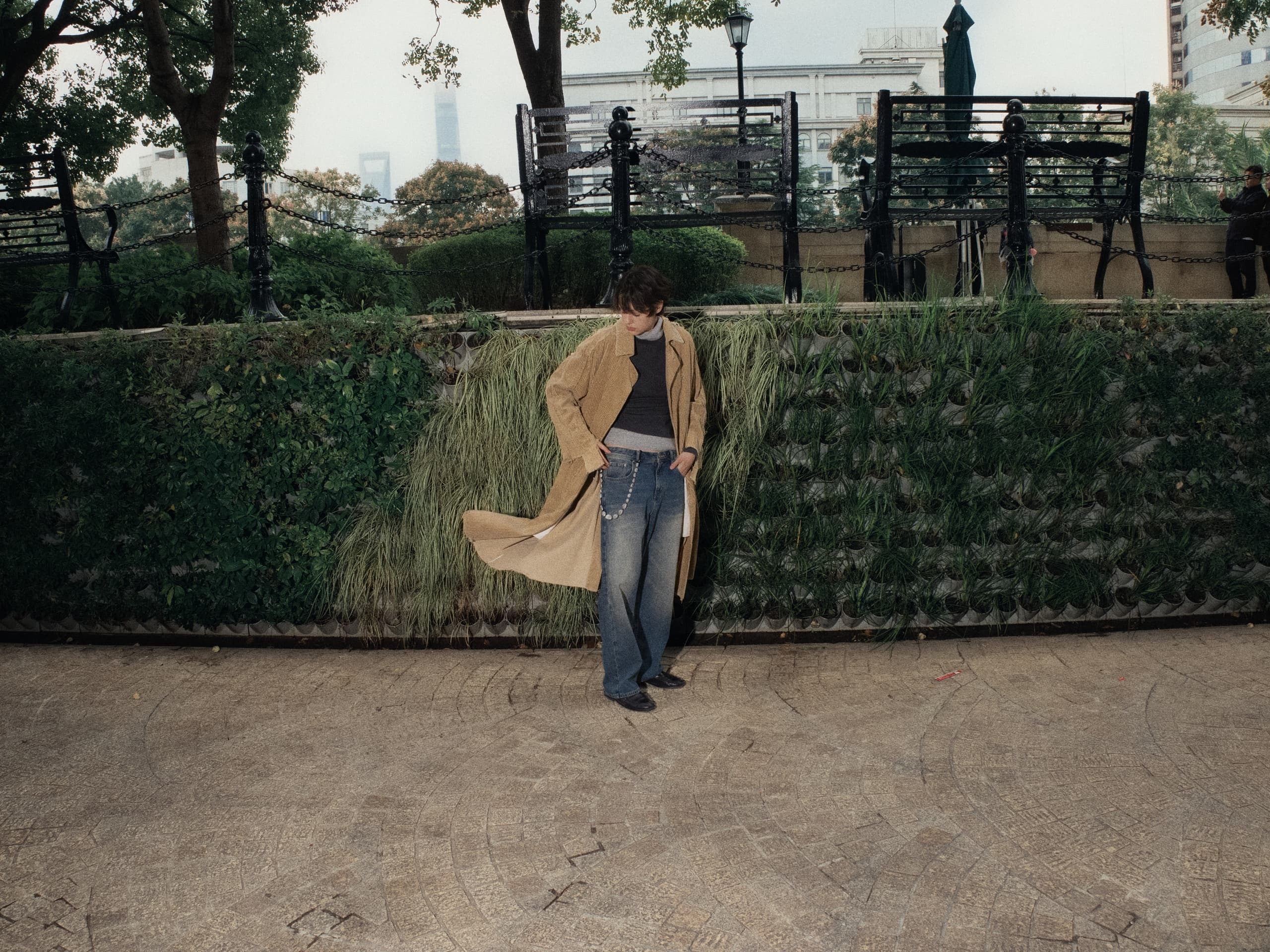 Person in a long coat poses by a green wall in a park setting.