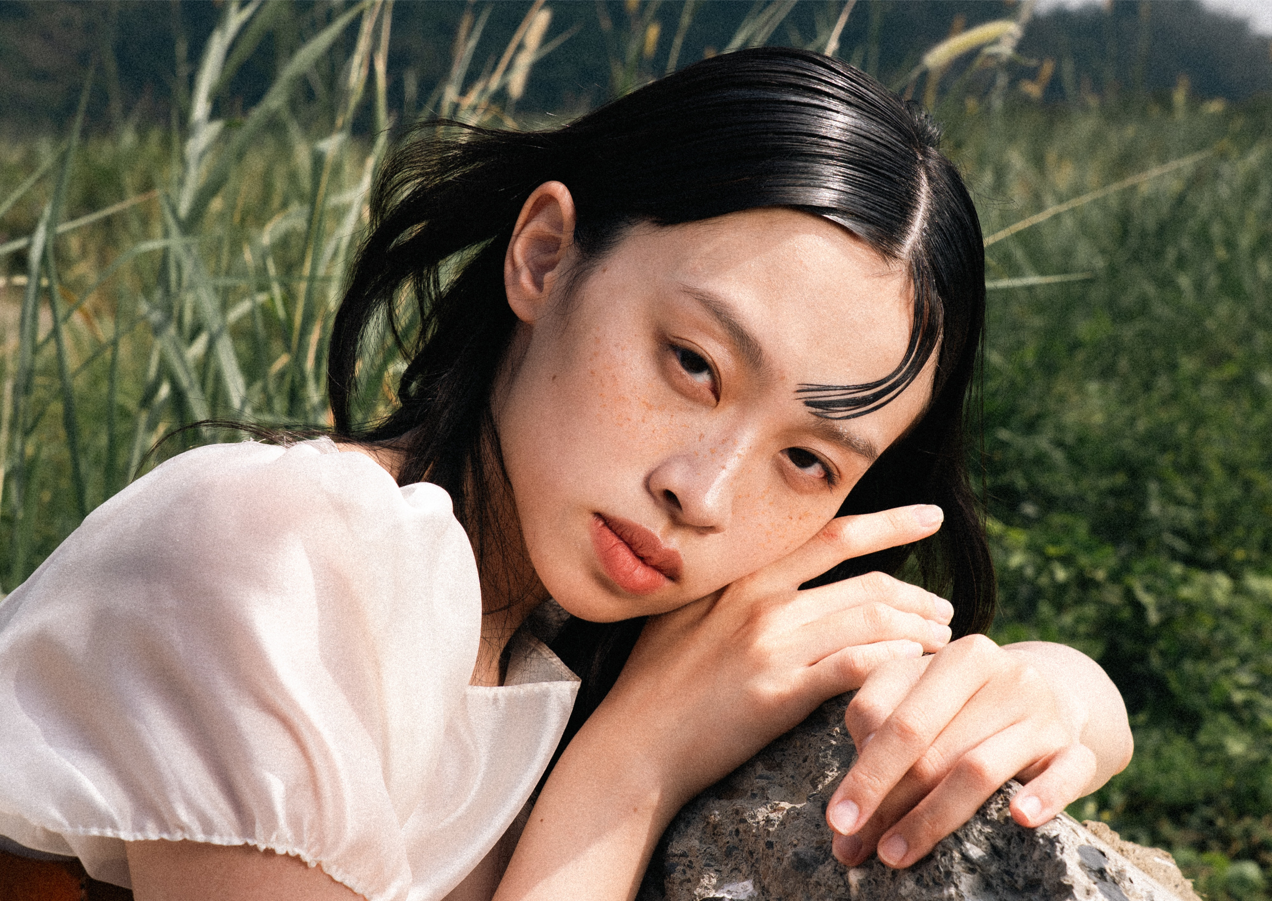 Young woman poses in nature with serene expression and delicate white blouse.