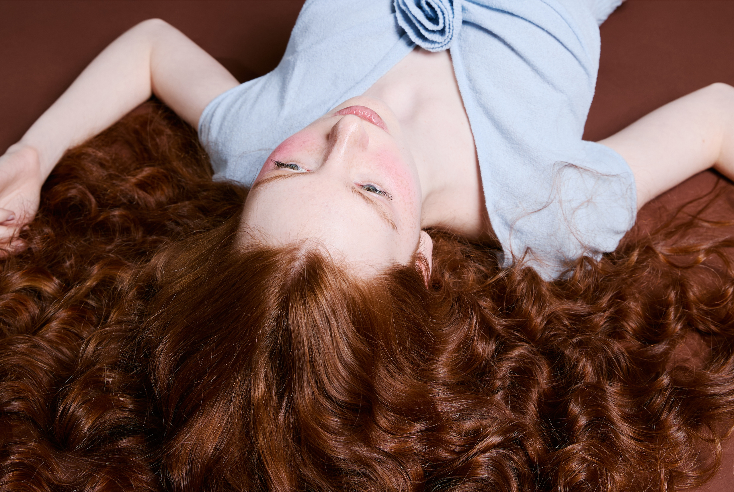 Woman with long curly red hair lying on a brown surface, wearing a light blue top.