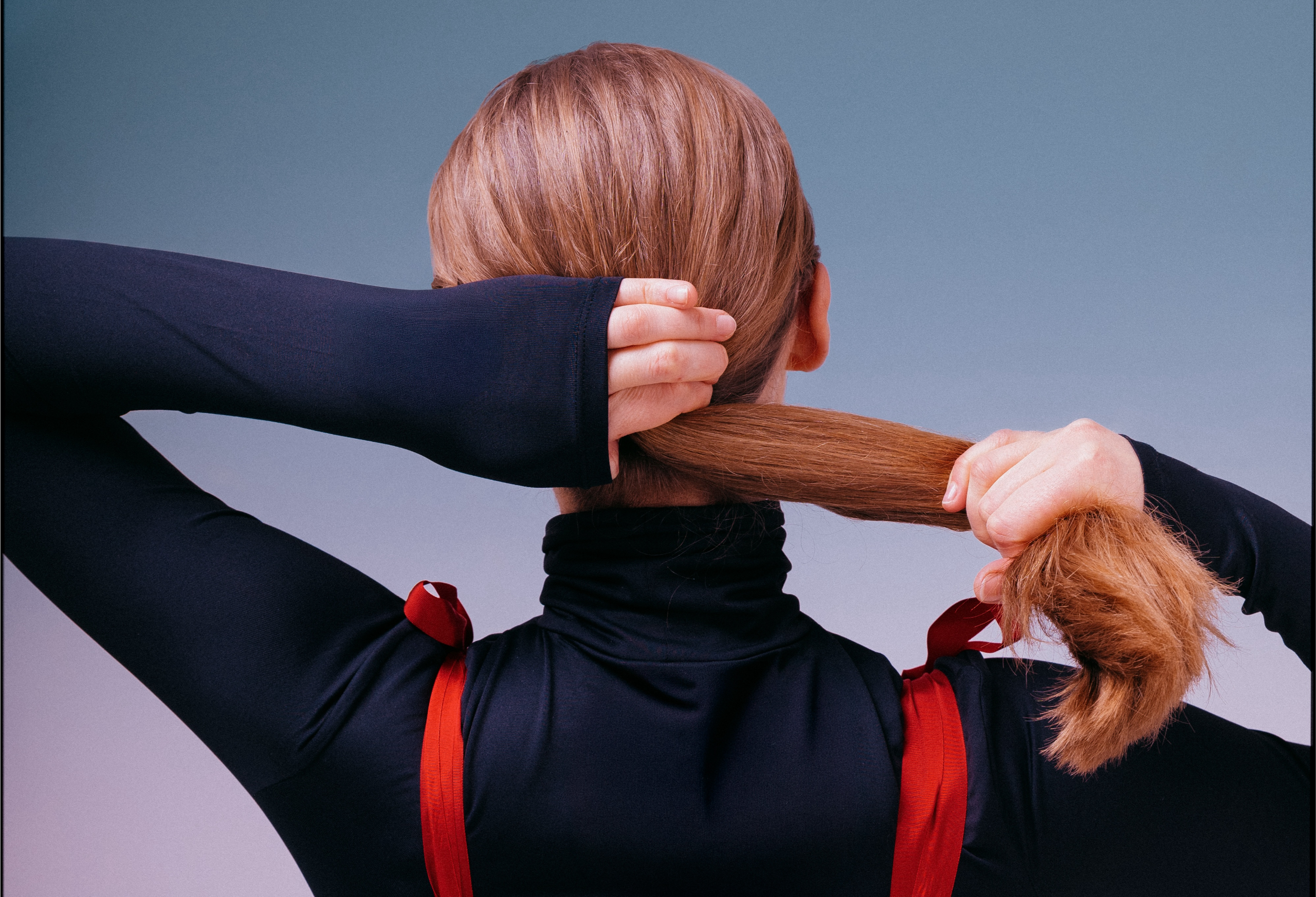 Person tying long hair into a ponytail, wearing a black top with red straps.