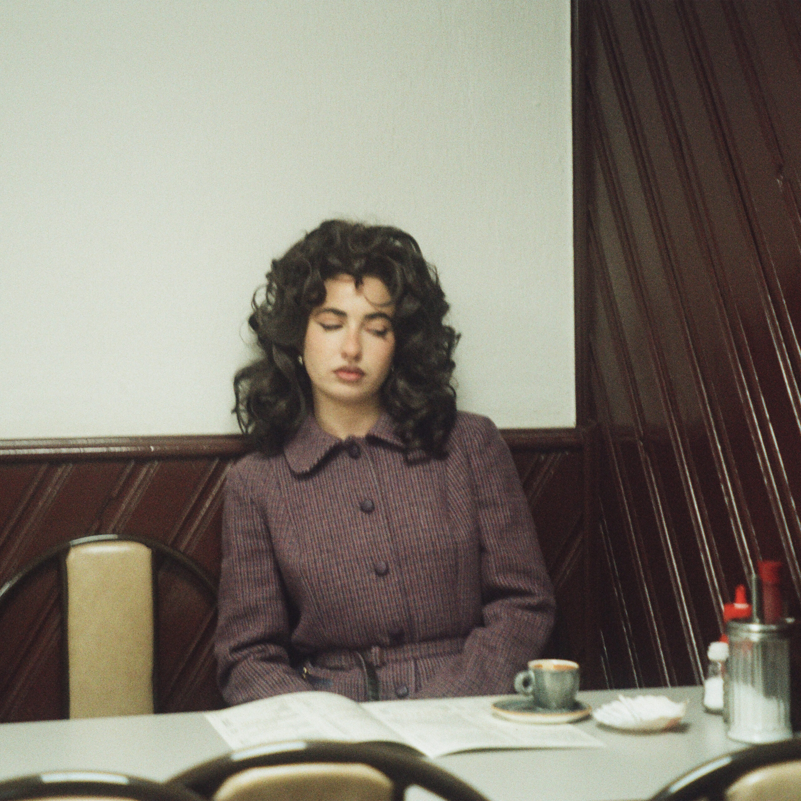 Woman in vintage coat sitting at a café table with a closed newspaper and coffee.