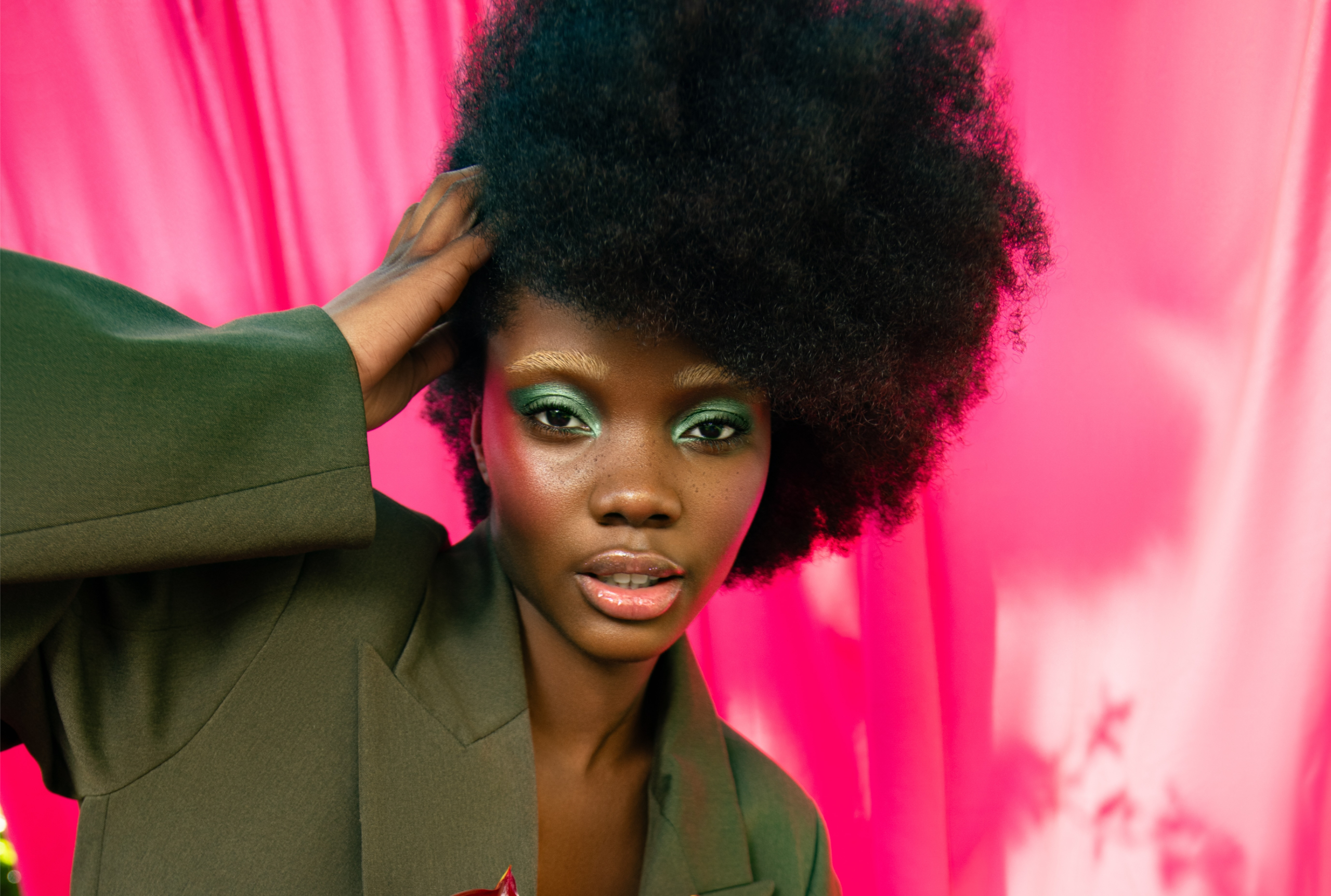 Model poses with vibrant makeup and afro hairstyle against a pink backdrop.