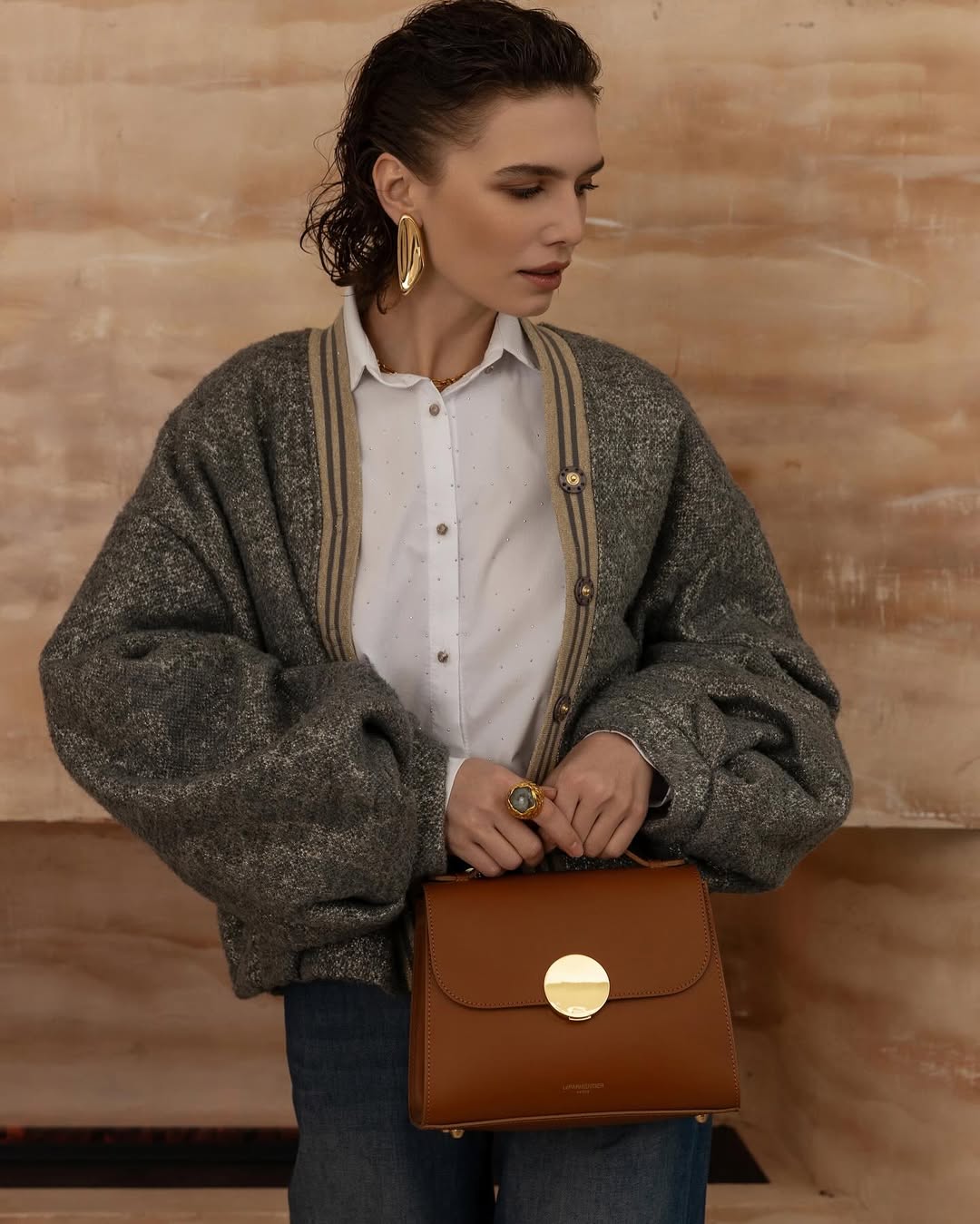 Woman in stylish cardigan holding a brown handbag with gold accents.