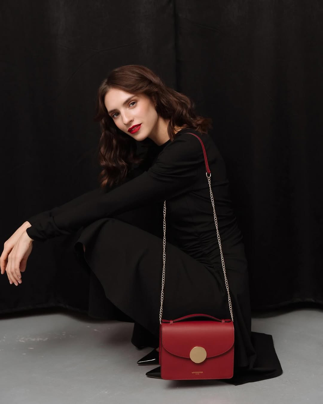 Woman in black with red handbag, sitting gracefully against a dark background.