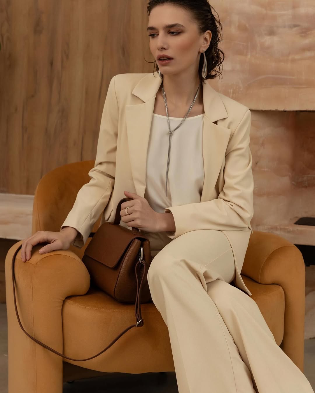 Woman in elegant beige suit sitting on a chair with a brown leather handbag.