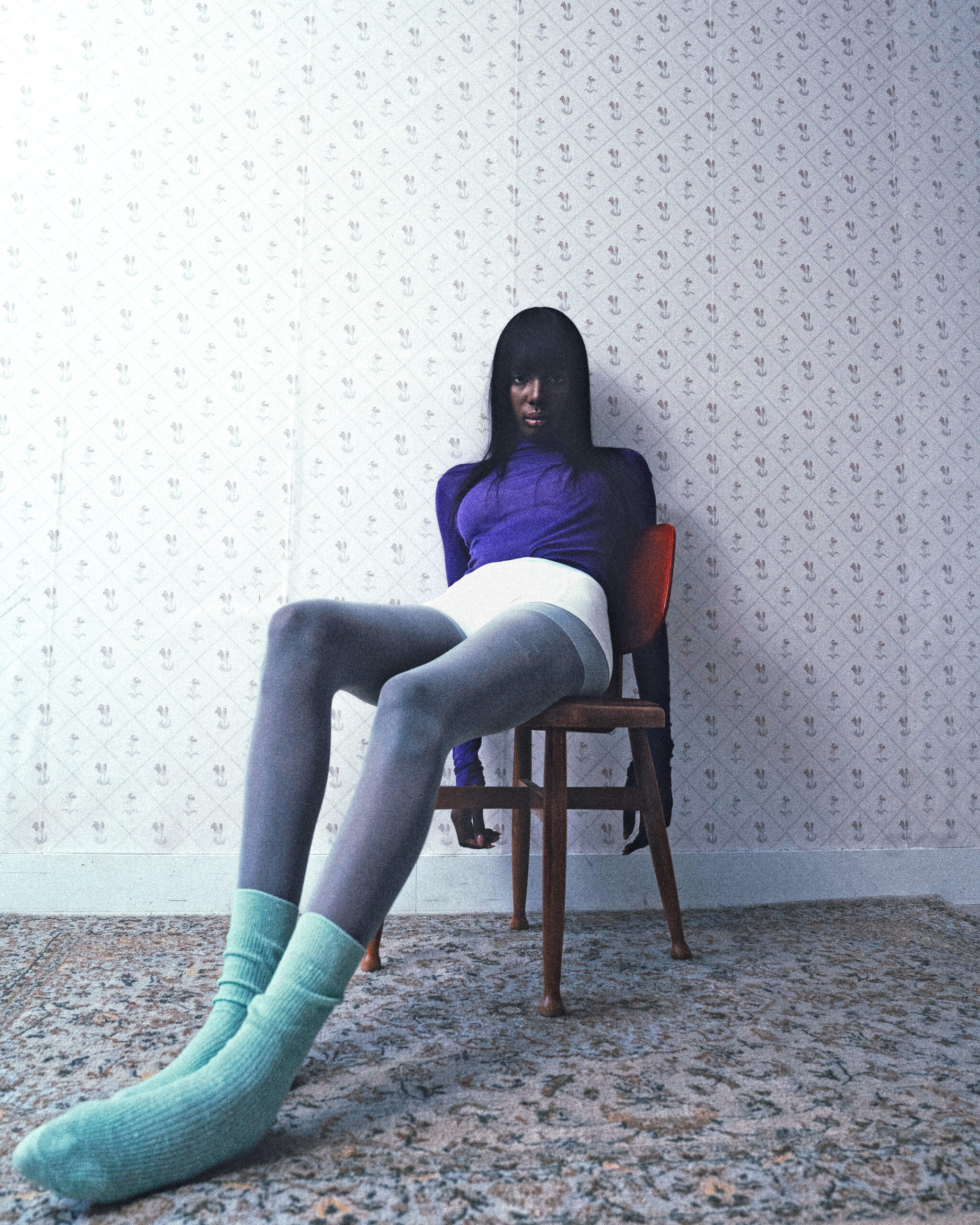 Person in colorful outfit sitting slouched on chair, patterned room background.