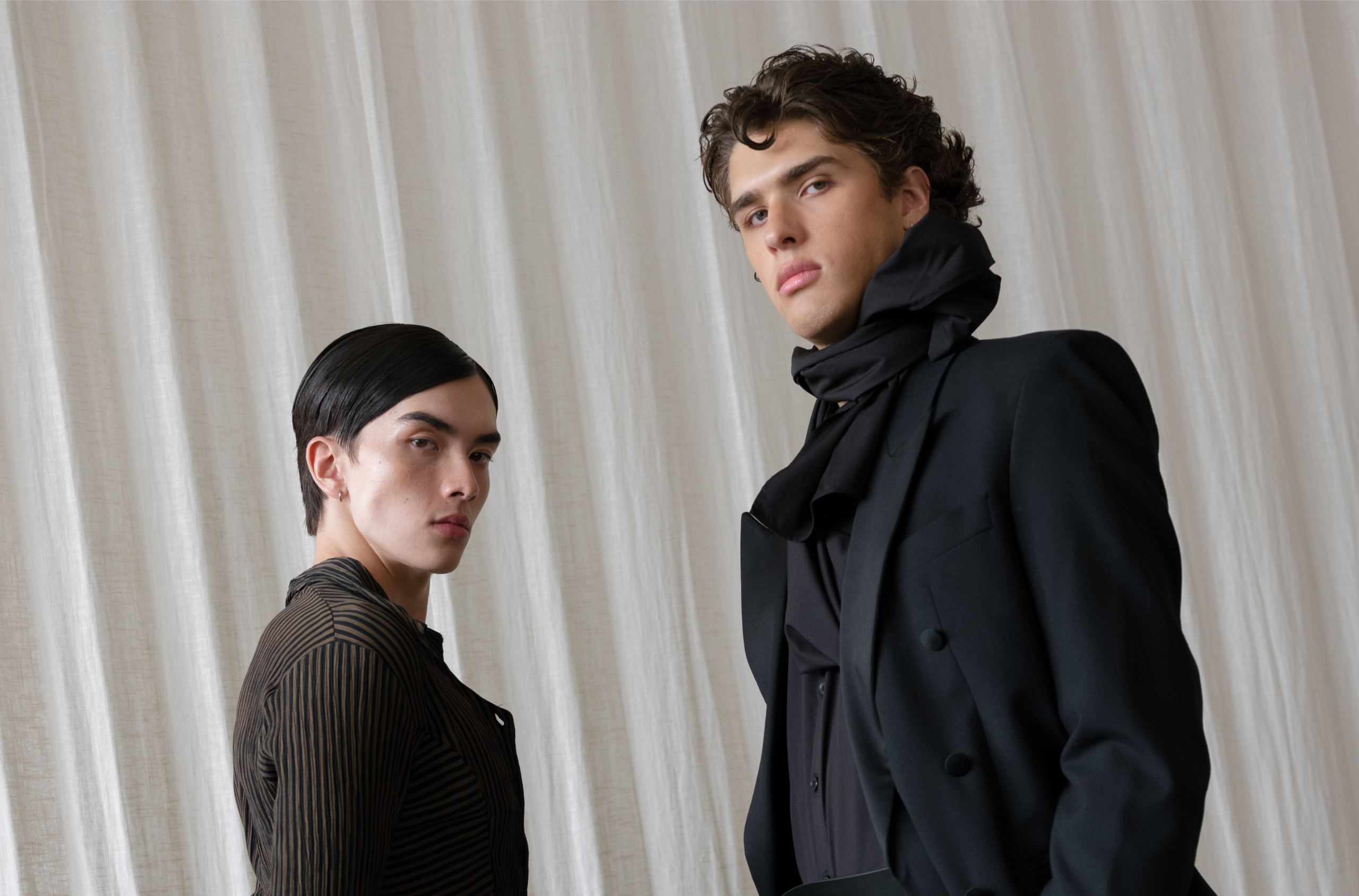 Two models in elegant fashion against a white curtain backdrop.