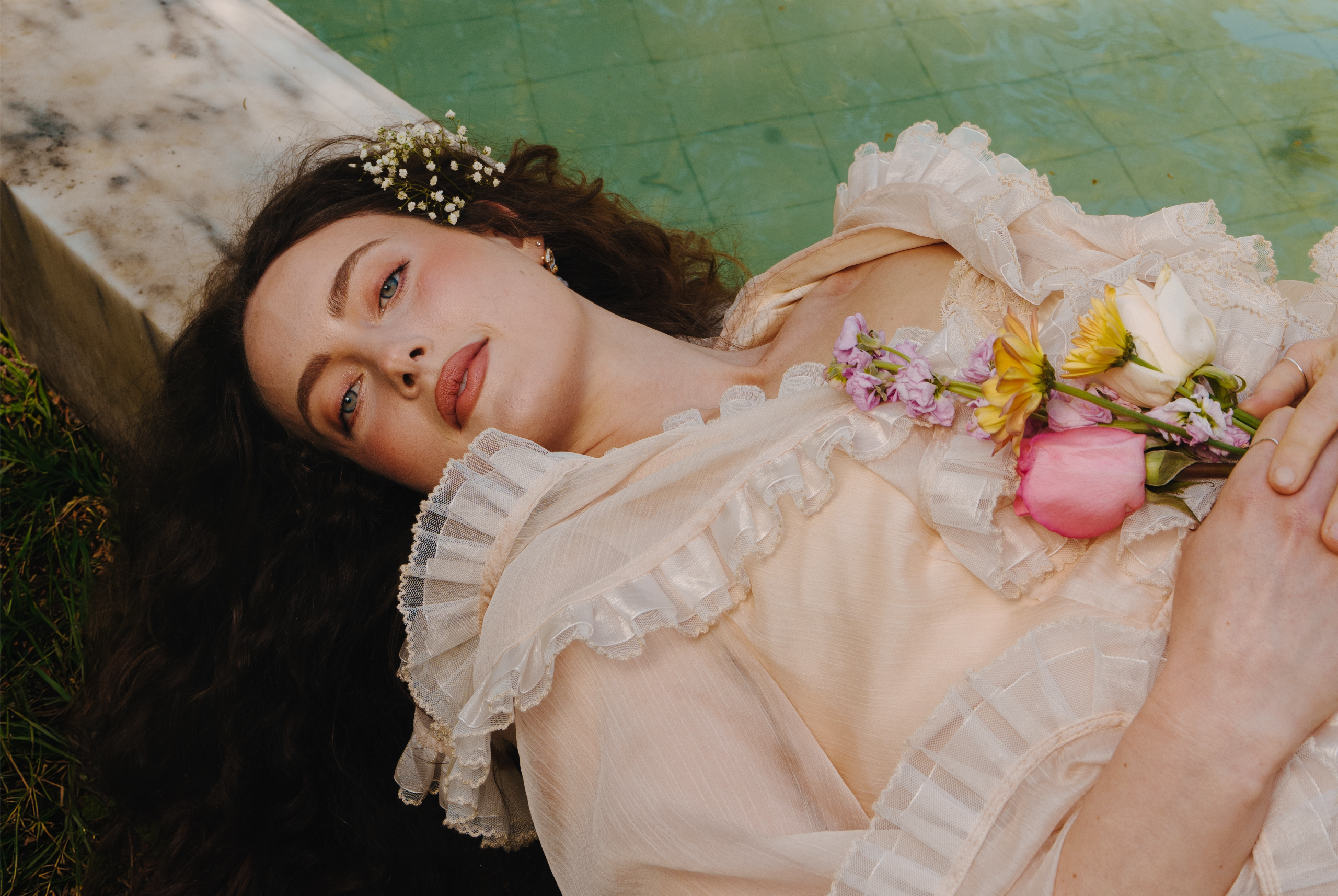 Woman in vintage dress lying by pool, holding colorful flowers, serene expression.