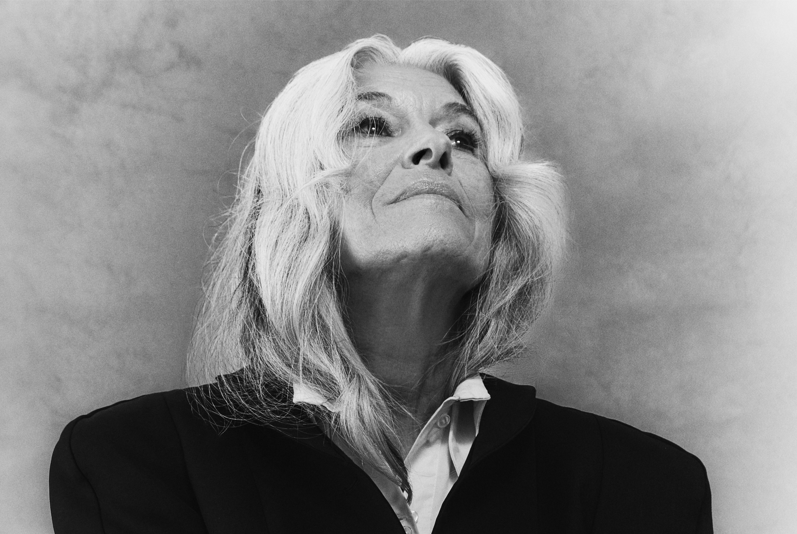 Elderly woman with long gray hair gazing upward in a suit, black and white photo.