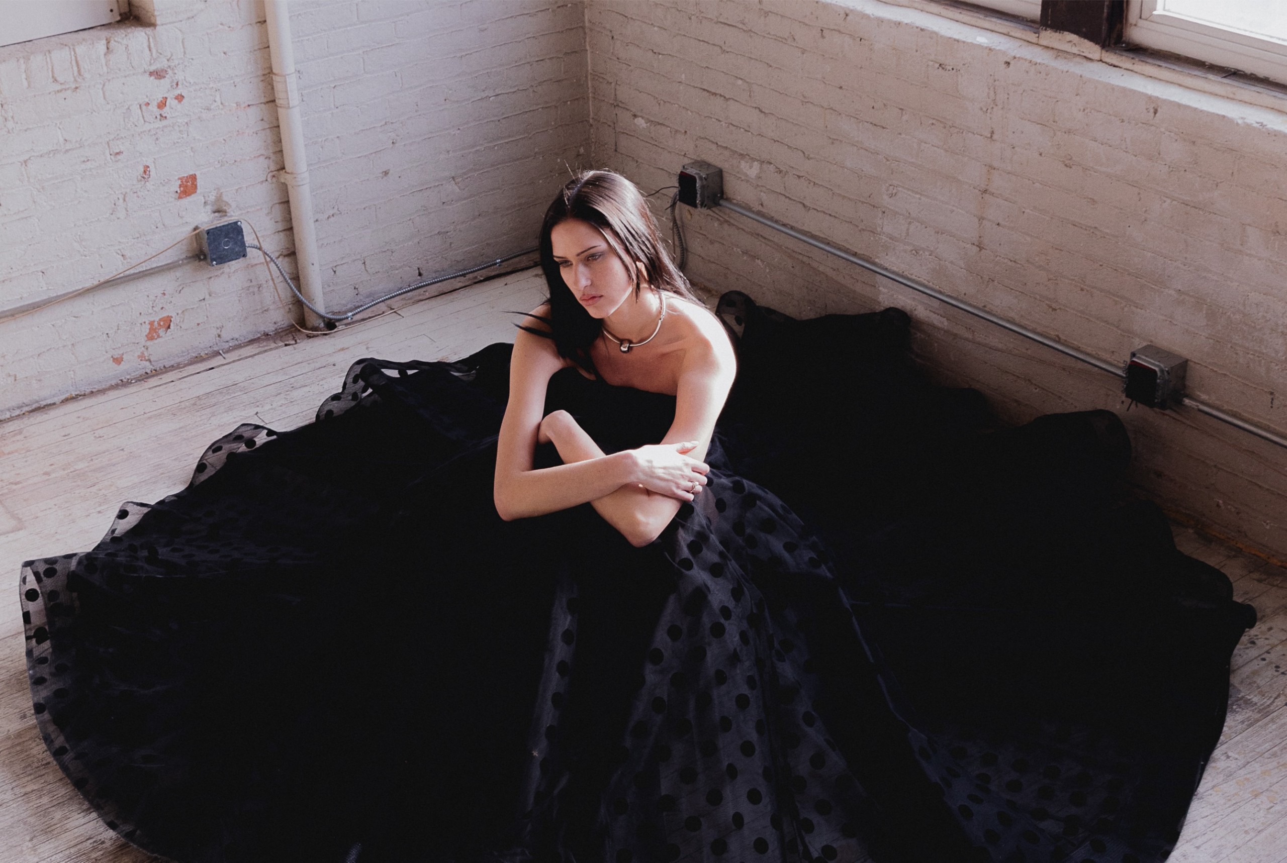 Woman in elegant black polka dot gown sitting in sunlit loft room.
