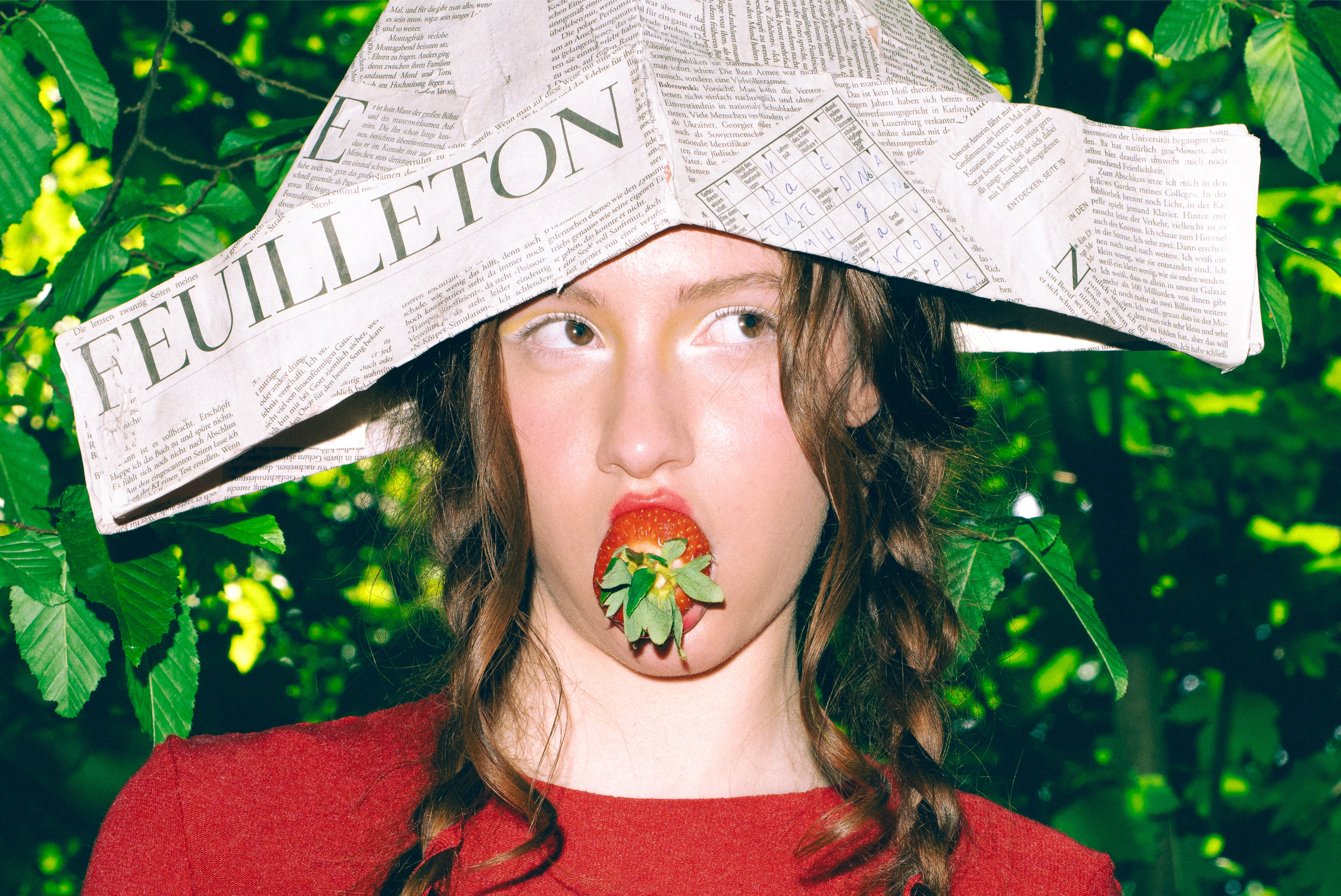 Woman with newspaper hat and strawberry in mouth, lush green background.