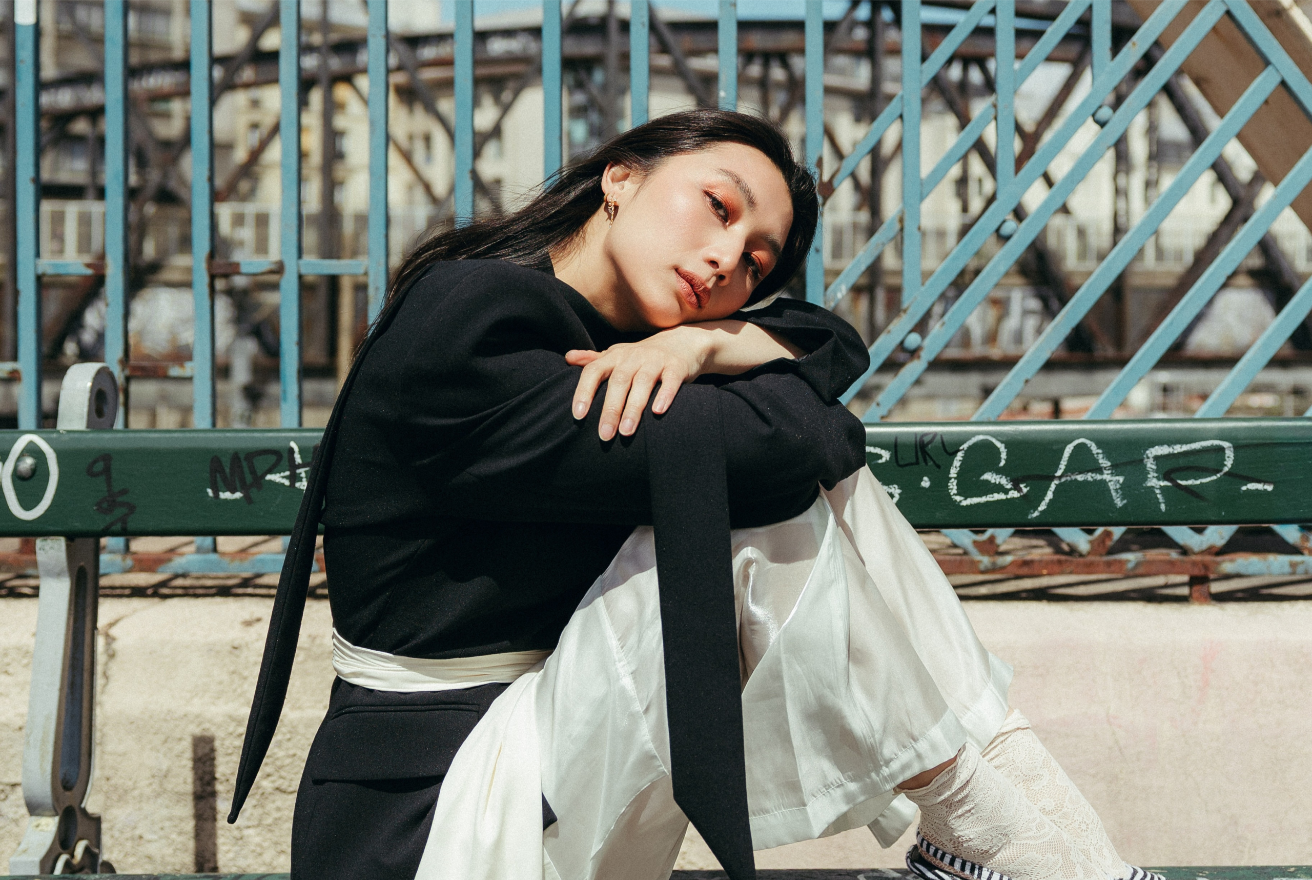 Fashionable person in urban setting, black jacket, white pants, sitting on bench.