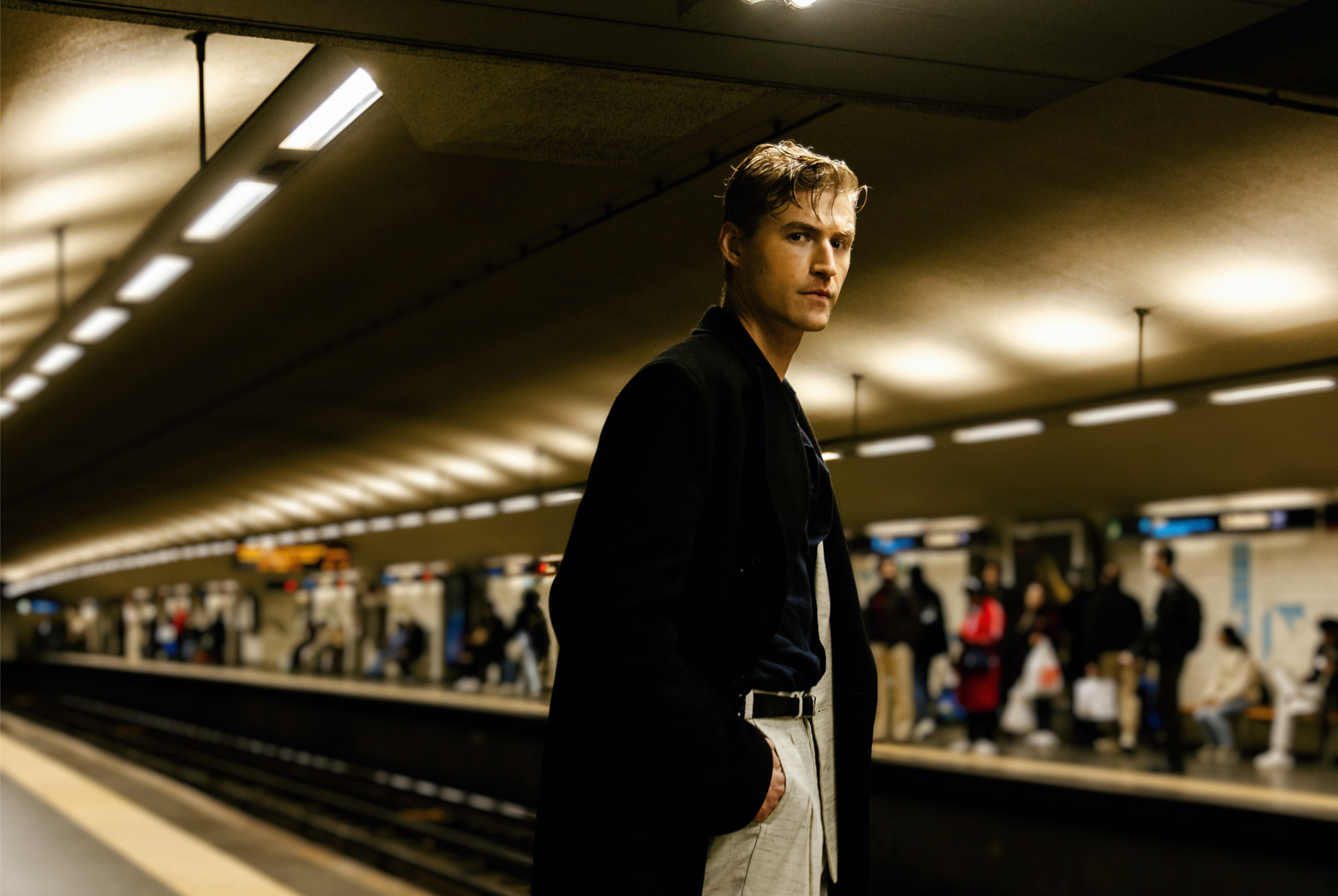 Man in a subway station wearing a dark coat with people waiting on the platform.