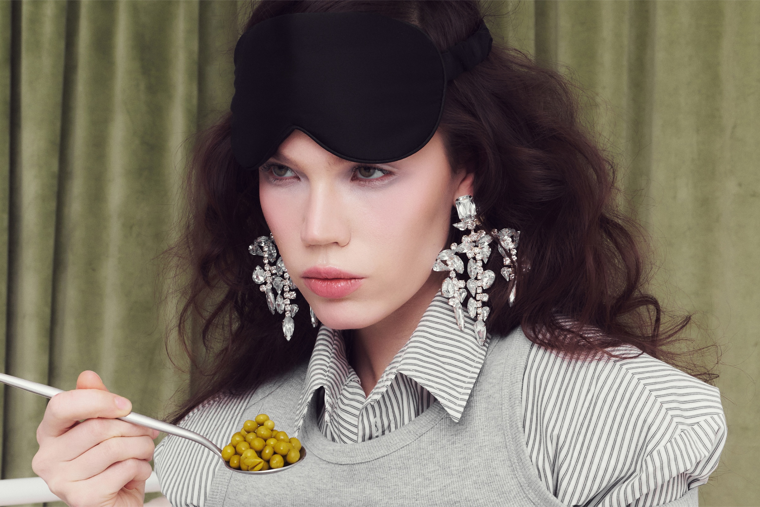 Fashionable woman with eye mask, eating peas, wearing large crystal earrings.