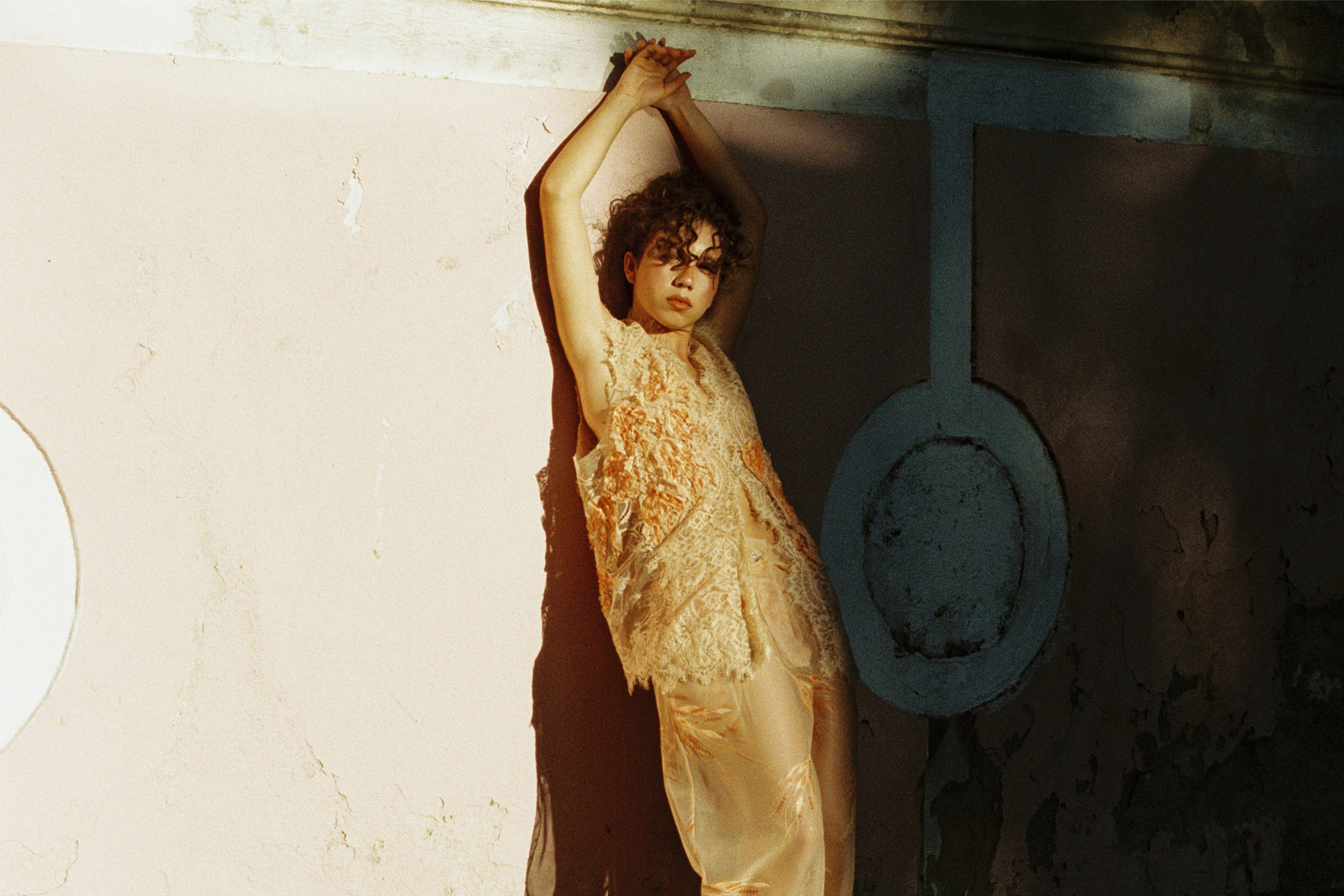 Person posing in stylish peach outfit against a textured wall with shadows.