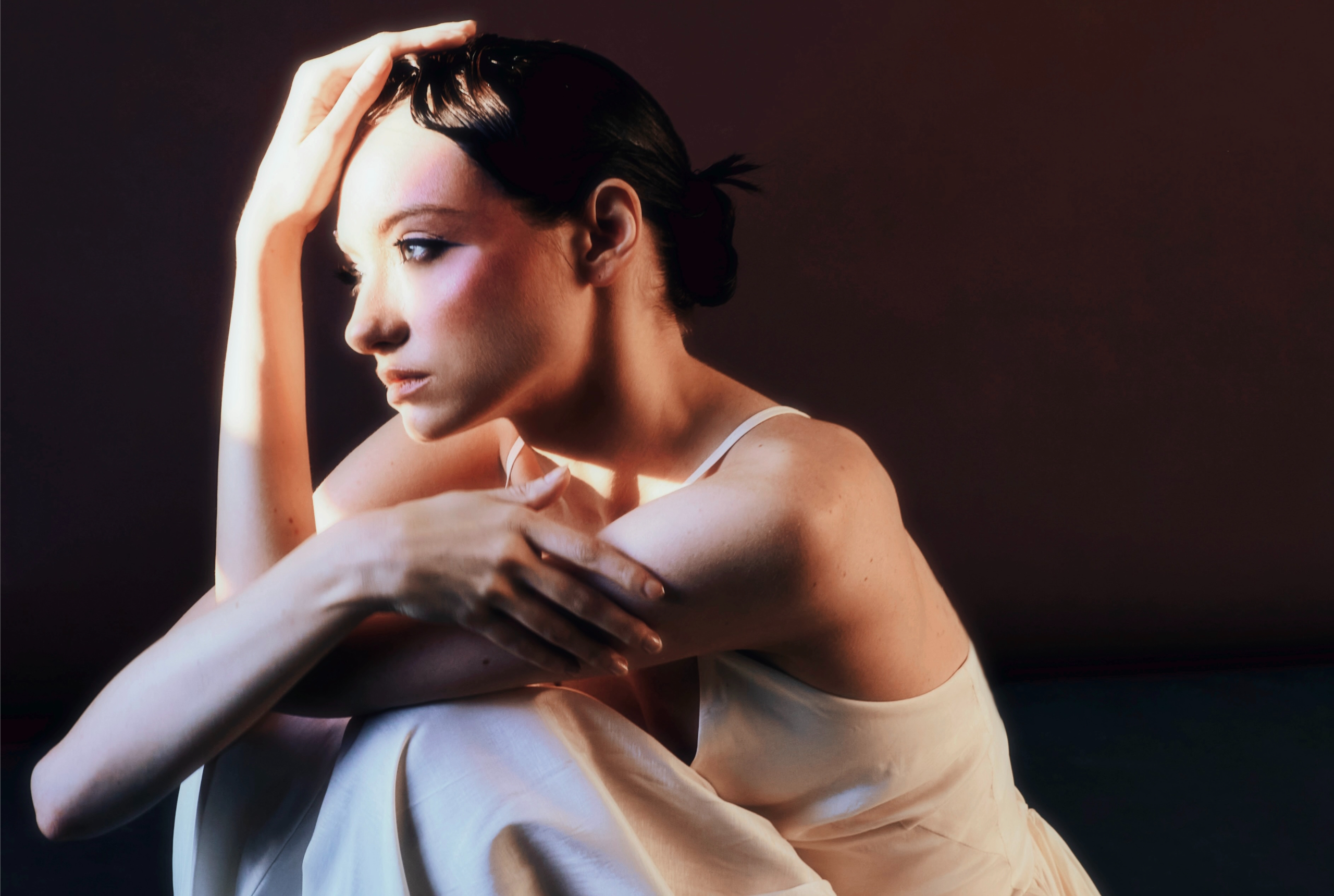 Woman in contemplative pose, soft light, wearing white dress, dark backdrop.