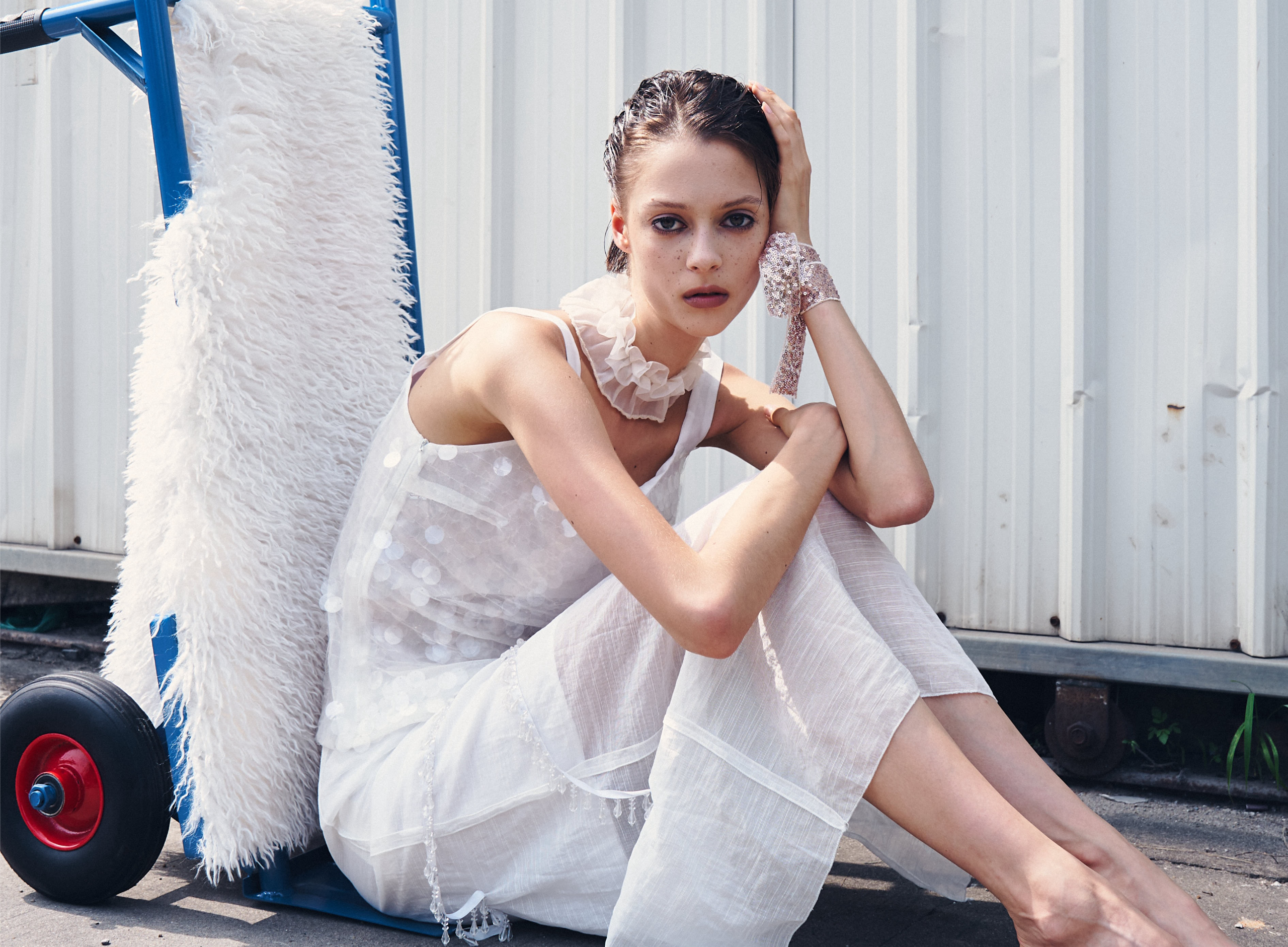 Woman in sheer white dress sits pensively by a cart in urban setting.