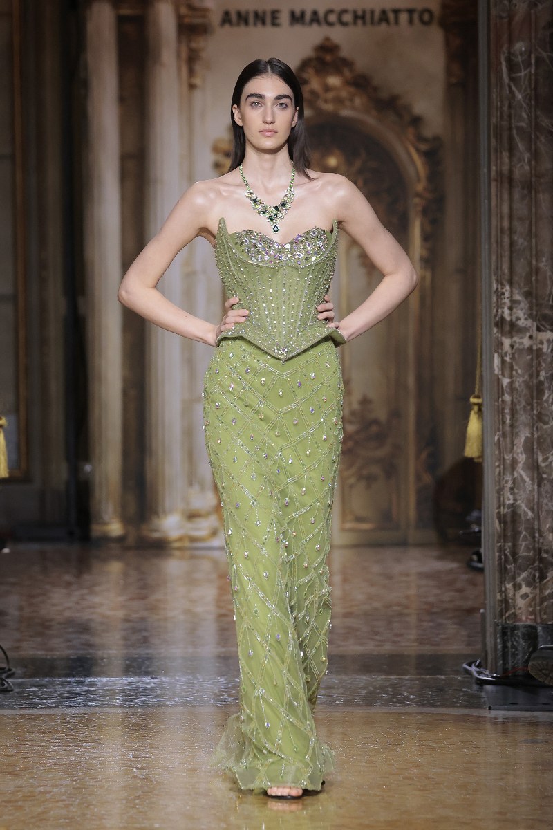 Model in an elegant green gown with ornate detailing on a fashion runway.