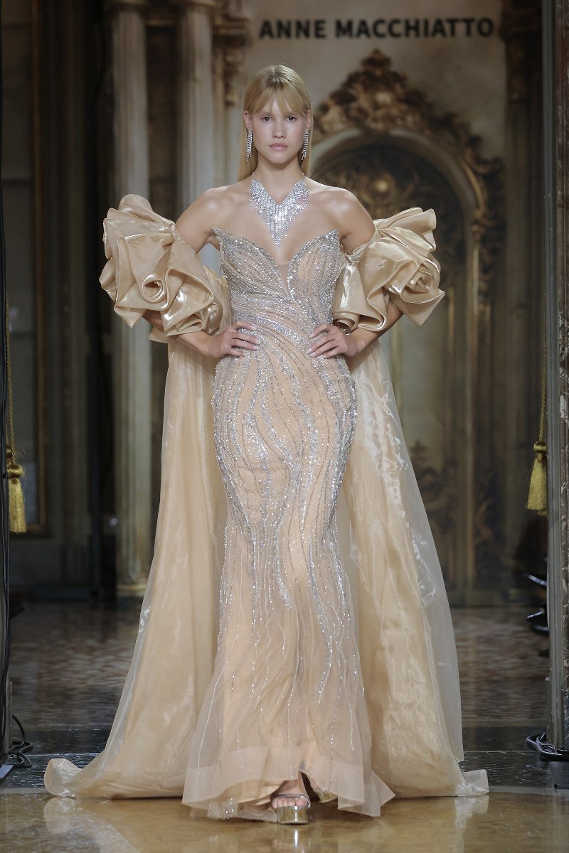 Model in elegant beige gown with bold sleeves on a runway.