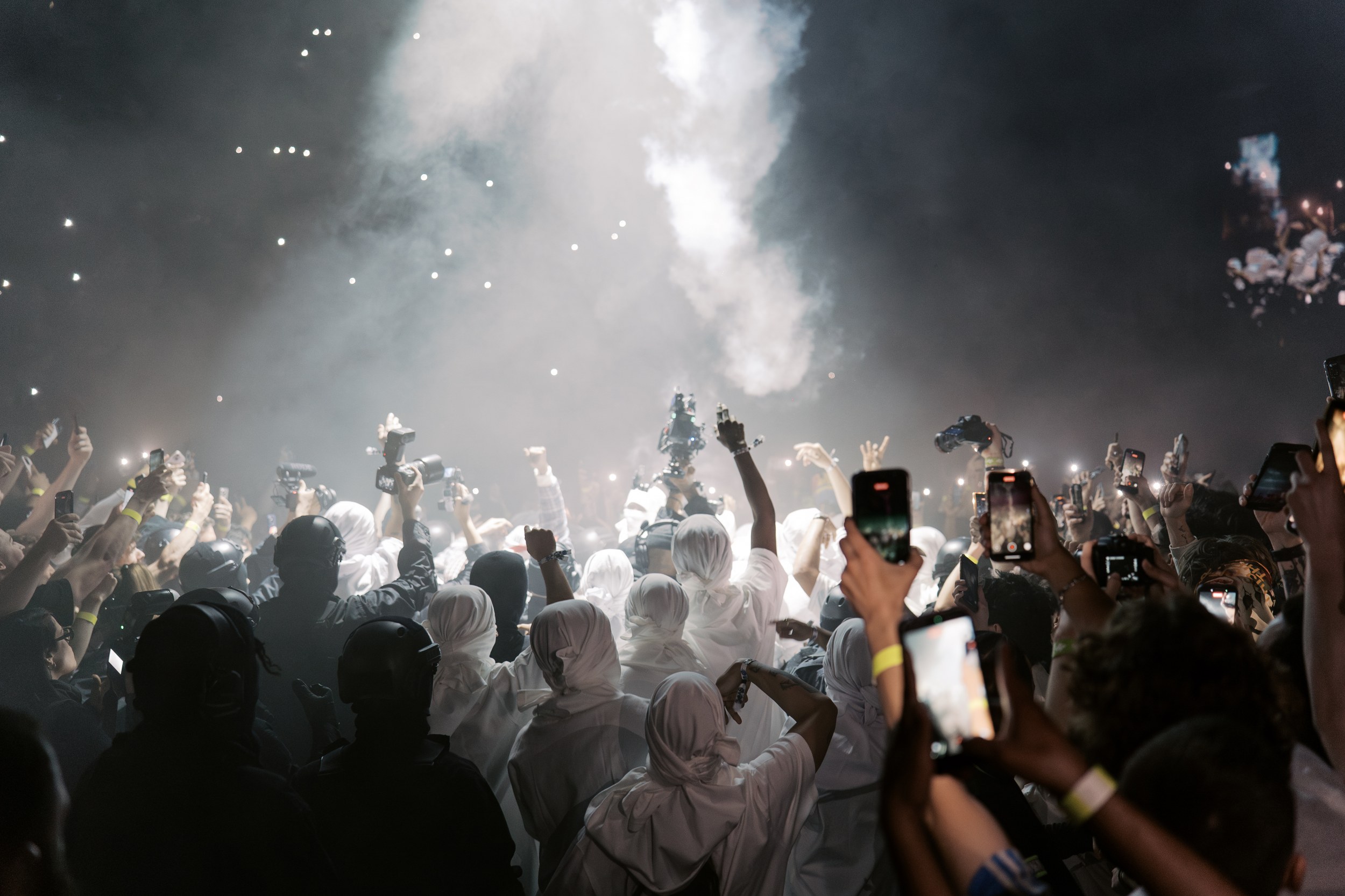 Concert crowd recording with phones, surrounded by smoke and lights.