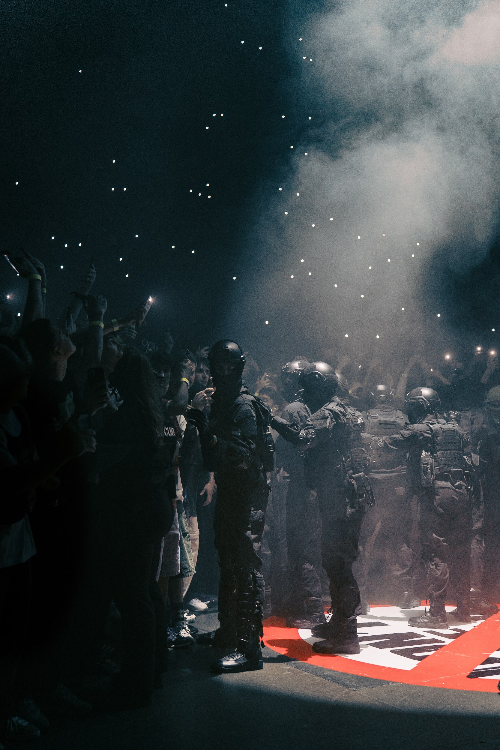 Riot police in dark, smoky atmosphere with crowd taking photos on phones.