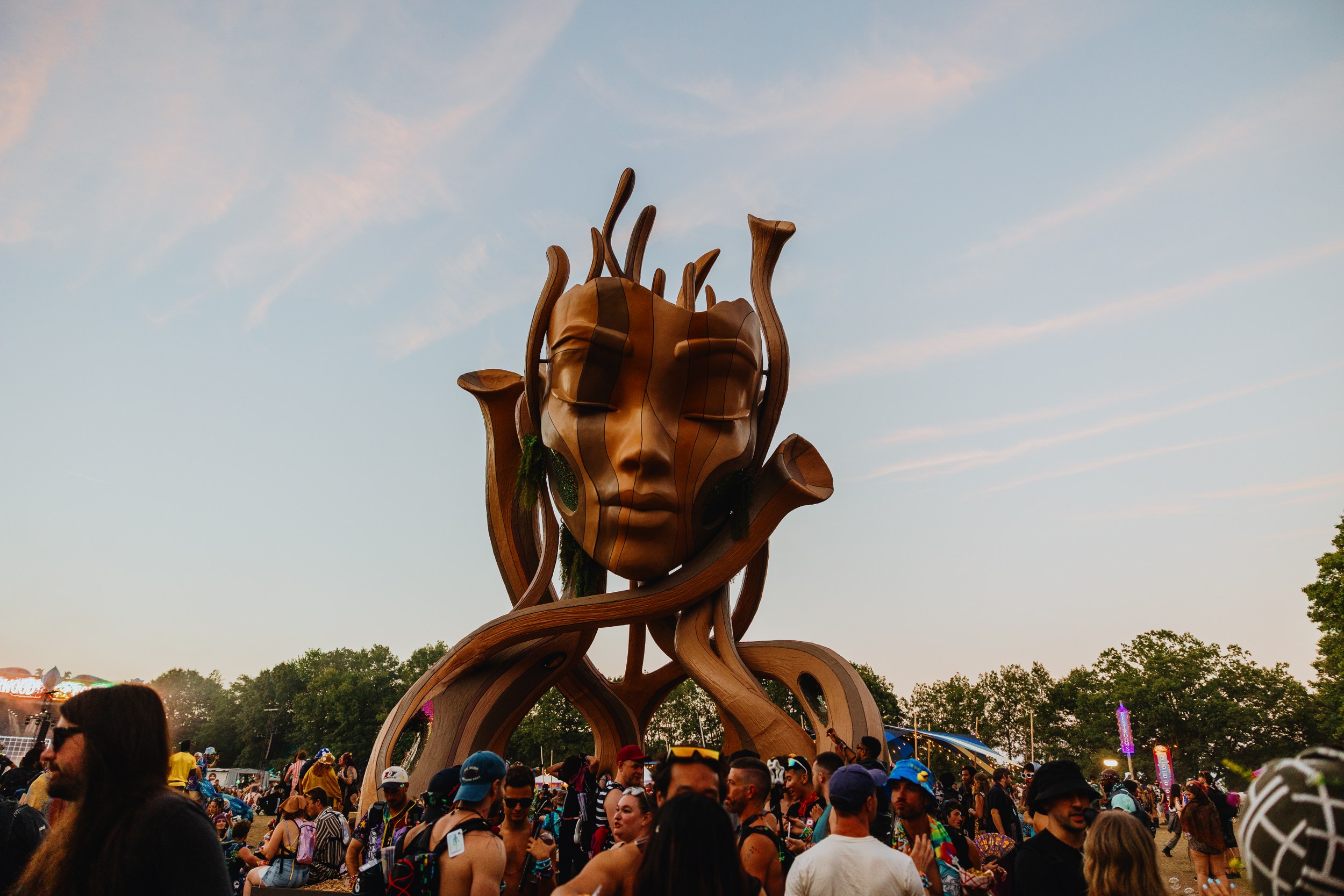 Festival crowd around a giant artistic wooden sculpture under a colorful sky.