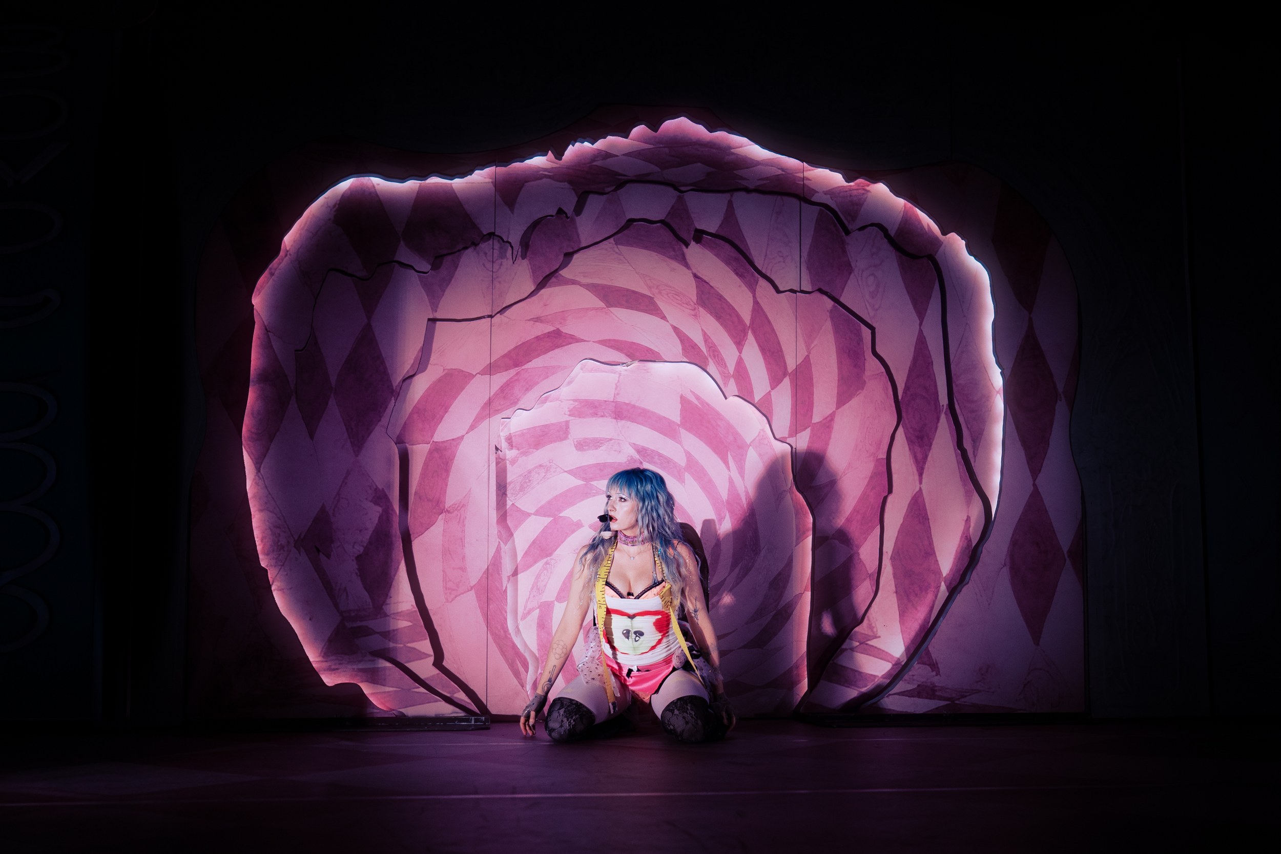 Performer kneels in front of a vibrant, illuminated pink spiral backdrop.